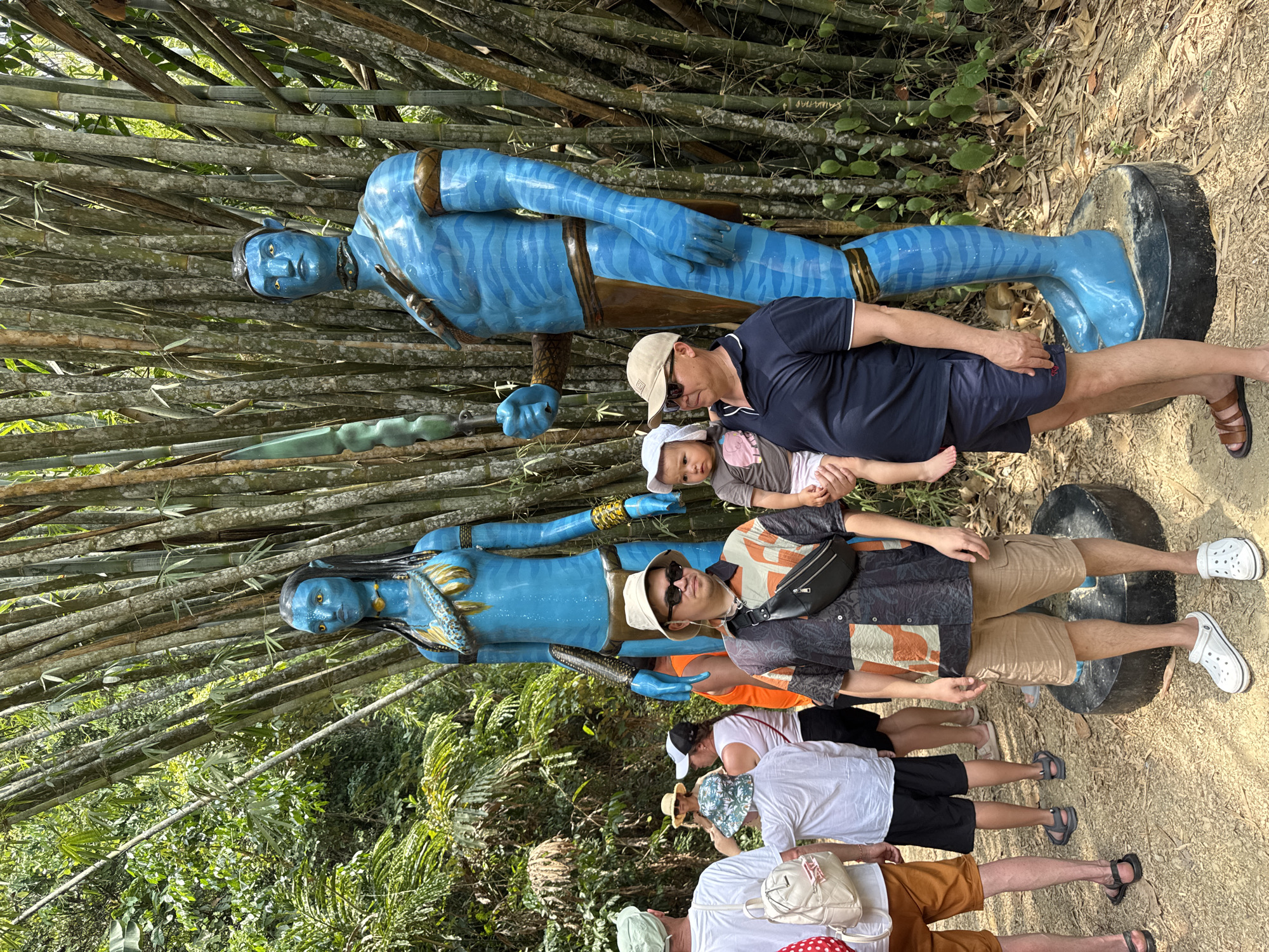 The image captures a group of people posing with statues of characters from the movie "Avatar" in Ban Khlong Khian, Thailand. The statues are blue with detailed markings, one male and one female, positioned against a bamboo backdrop. A man in a dark shirt and hat is holding a baby, while another man in a patterned shirt and sunglasses stands next to them. The other members of the group are standing in line behind them. The environment consists of lush greenery and a dirt path.