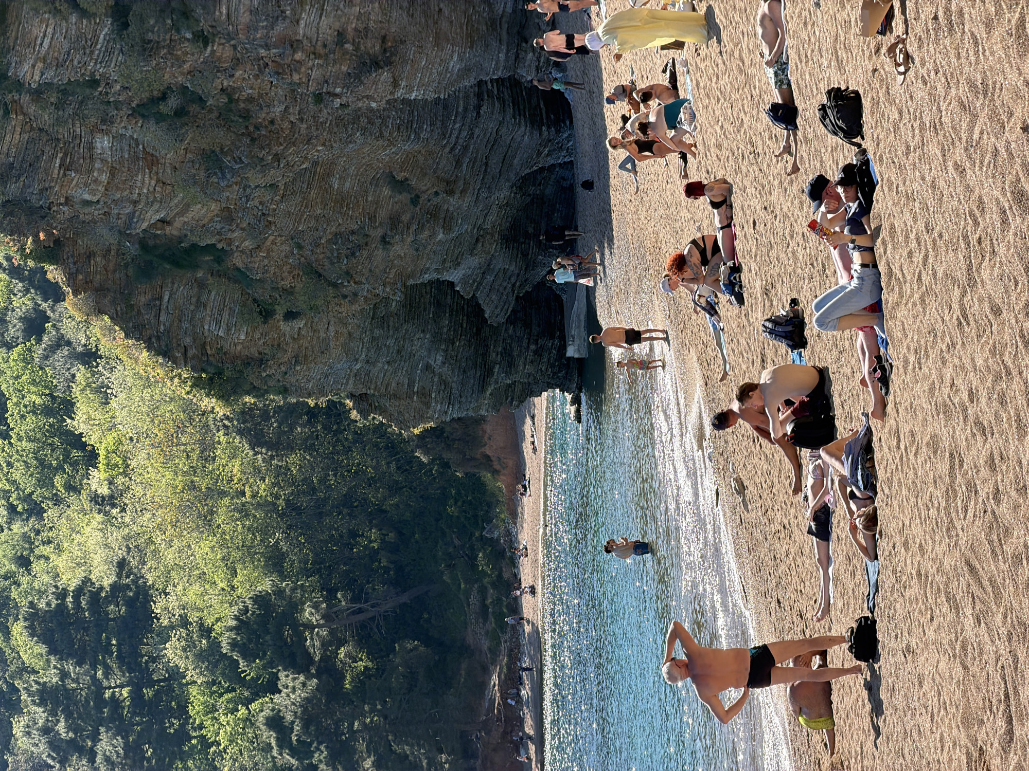 The image captures a bustling scene at Mogren Beach in Budva, Montenegro. The beach is nestled between rocky cliffs covered in lush greenery and the sparkling Adriatic Sea. Numerous people are present: some sunbathing on the sandy shore, others swimming in the water, and some walking along the beach. The scene suggests a warm, sunny day.