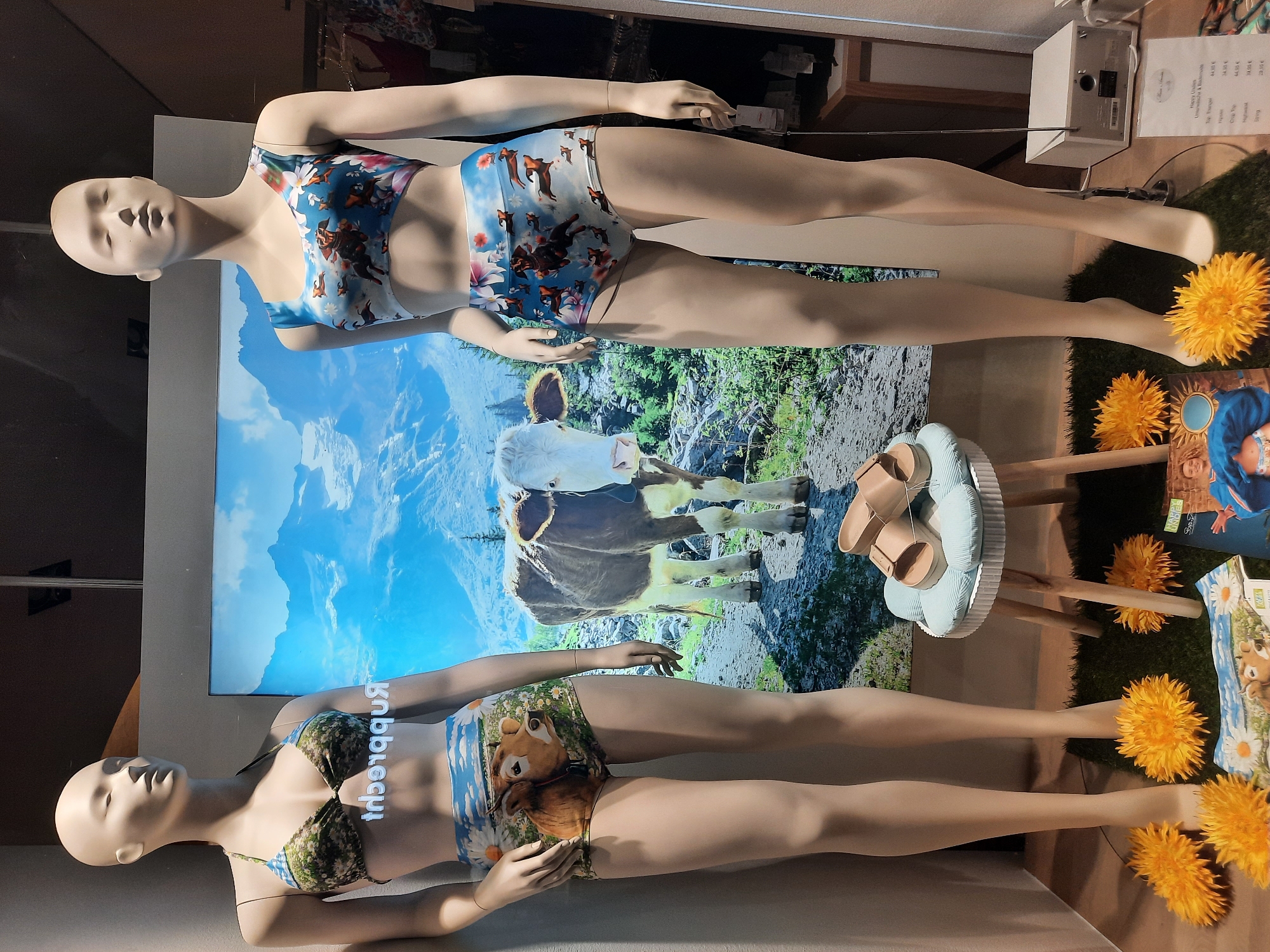 The image displays a store window in Rosenheim, Germany, featuring two mannequins dressed in swimwear. The mannequin on the left wears a camouflage bikini with daisy and cow print bottoms. The mannequin on the right wears a blue bikini top with dachshund print and matching bottoms. Behind the mannequins, a large backdrop depicts a mountainous landscape with cows. On a small stool between the mannequins are a pair of beige slides. The floor is covered with artificial grass, adorned with yellow flower decorations and a promotional item. A price list is visible to the right of the mannequin on the right.