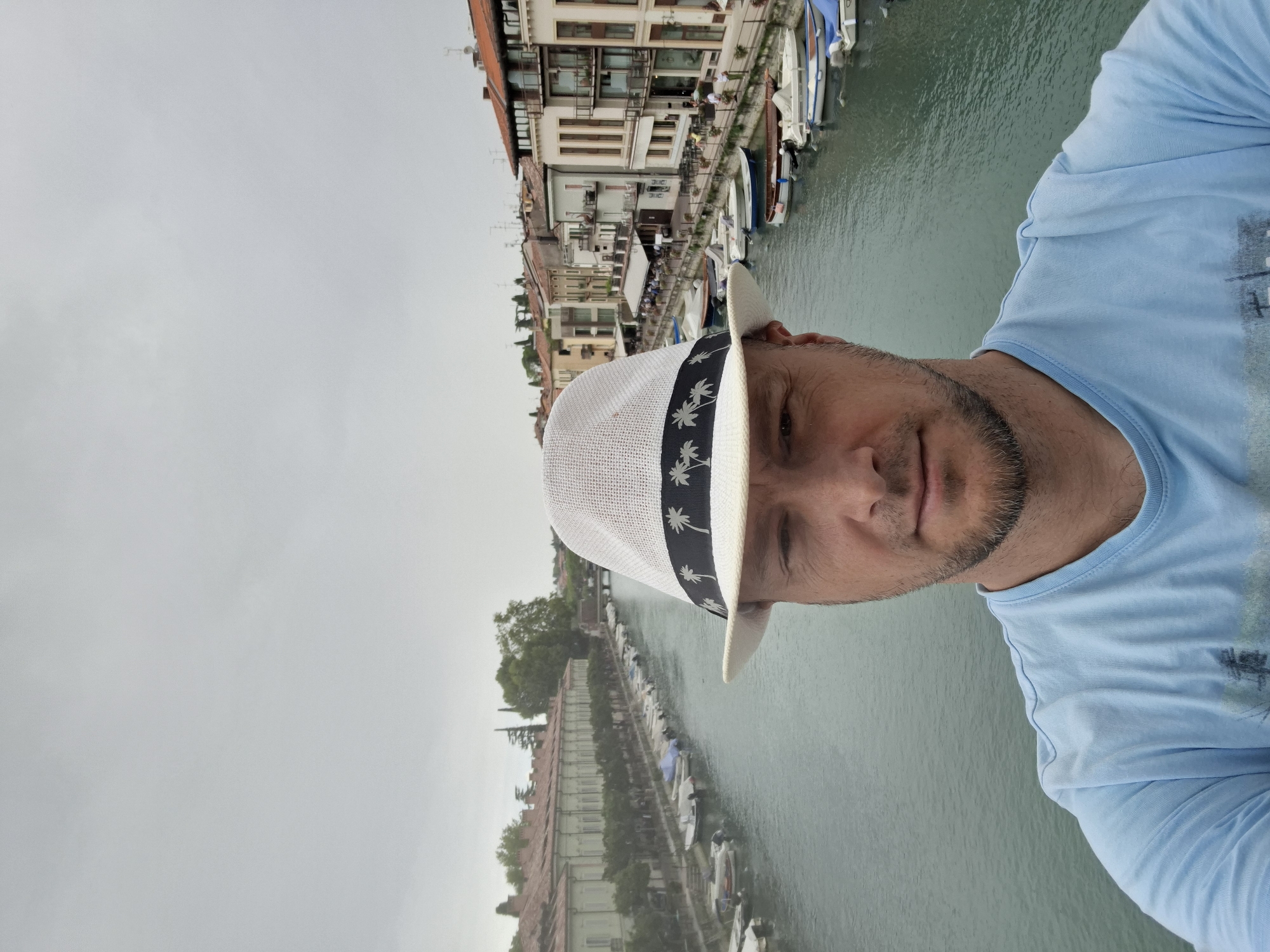 The image is a selfie taken by a man wearing a white hat with a black band decorated with palm trees, and a light blue t-shirt. He is looking directly at the camera. Behind him is a canal lined with boats, buildings with red-tiled roofs, and trees. The architecture and canal suggest a European city, consistent with Peschiera del Garda, Italy, which is known for its canals and Venetian fortifications. The sky is overcast and grey. The man's face is partially obscured by the brim of his hat.