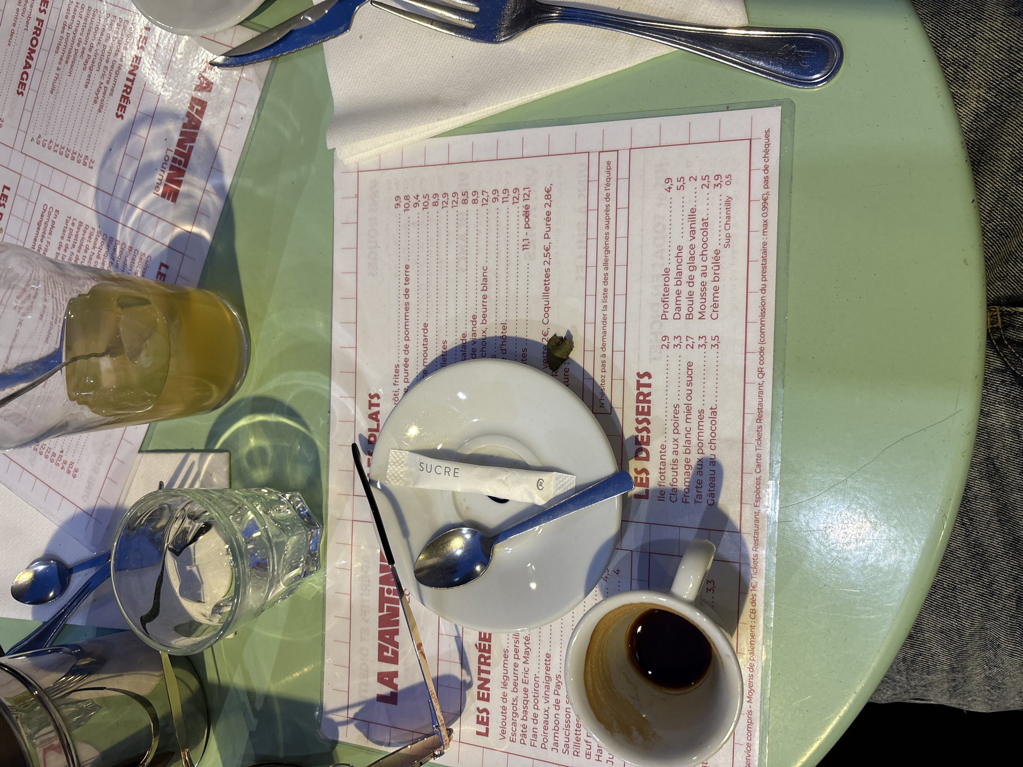 The image presents a close-up, top-down view of a dining table in a casual restaurant or "cantine" located in Paris, France. The light green table surface, showing visible scratches and wear, is primarily occupied by a laminated white menu with red text, titled "LA CANTINE," listing "LES ENTRÉES," "PLATS," and "LES DESSERTS" with prices in Euros.
Essential details include:
* **Objects on the table:** A pair of gold-framed reading glasses rests on the menu, beside a small white saucer containing a silver spoon and a white "SUCRE" packet. To the left, a white ceramic coffee cup holds residual dark liquid, consistent with espresso. A clear glass, partially filled with water, and another glass containing an amber liquid (likely juice) with ice cubes and a spoon, are positioned above the menu. A metallic carafe is partially visible in the upper left corner. On the right side of the menu, a folded white napkin is placed, holding a silver four-tined fork and a knife with a blue handle.
* **Environment:** The setting is indoors, with bright lighting casting distinct shadows of the objects onto the table and menu.
* **Actions/Interactions:** The arrangement of items, including partially consumed beverages and cutlery, indicates a recent or ongoing meal. No individuals are visible, but the scene implies human activity.