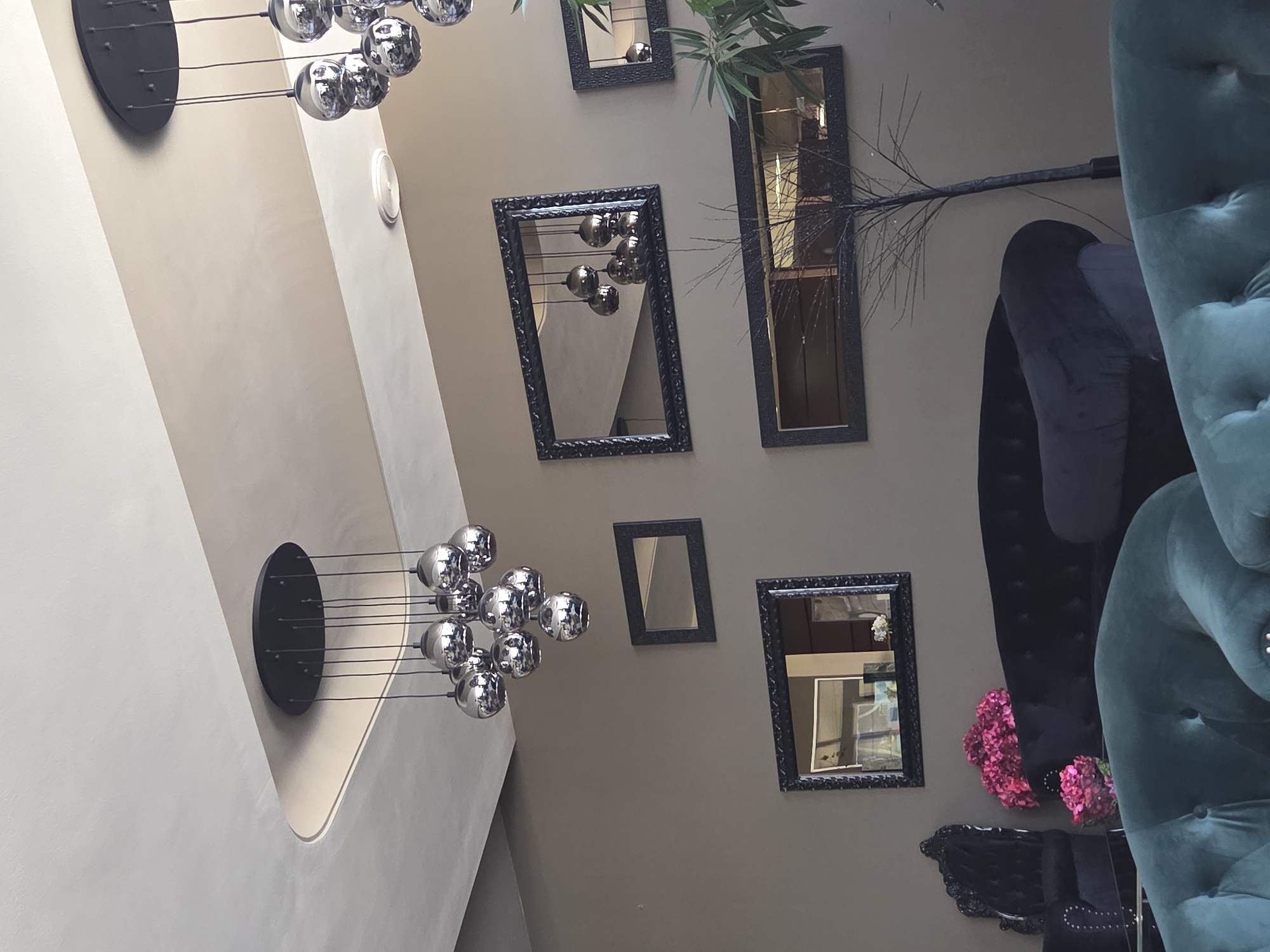 The image shows an interior setting with a combination of modern and ornate decorative elements.
The background is a neutral-toned wall, possibly a beige or light brown, adorned with an assortment of framed mirrors of various sizes and shapes. Some mirrors reflect parts of the room.
A modern chandelier with multiple metallic spheres hangs from the ceiling.
In the foreground, partially visible, are plush, button-tufted velvet chairs in shades of teal and blue. There is a black ornate accent chair visible with two bouquets of pink flowers.
The exact location in Pleven, Bulgaria, cannot be determined from the image alone.