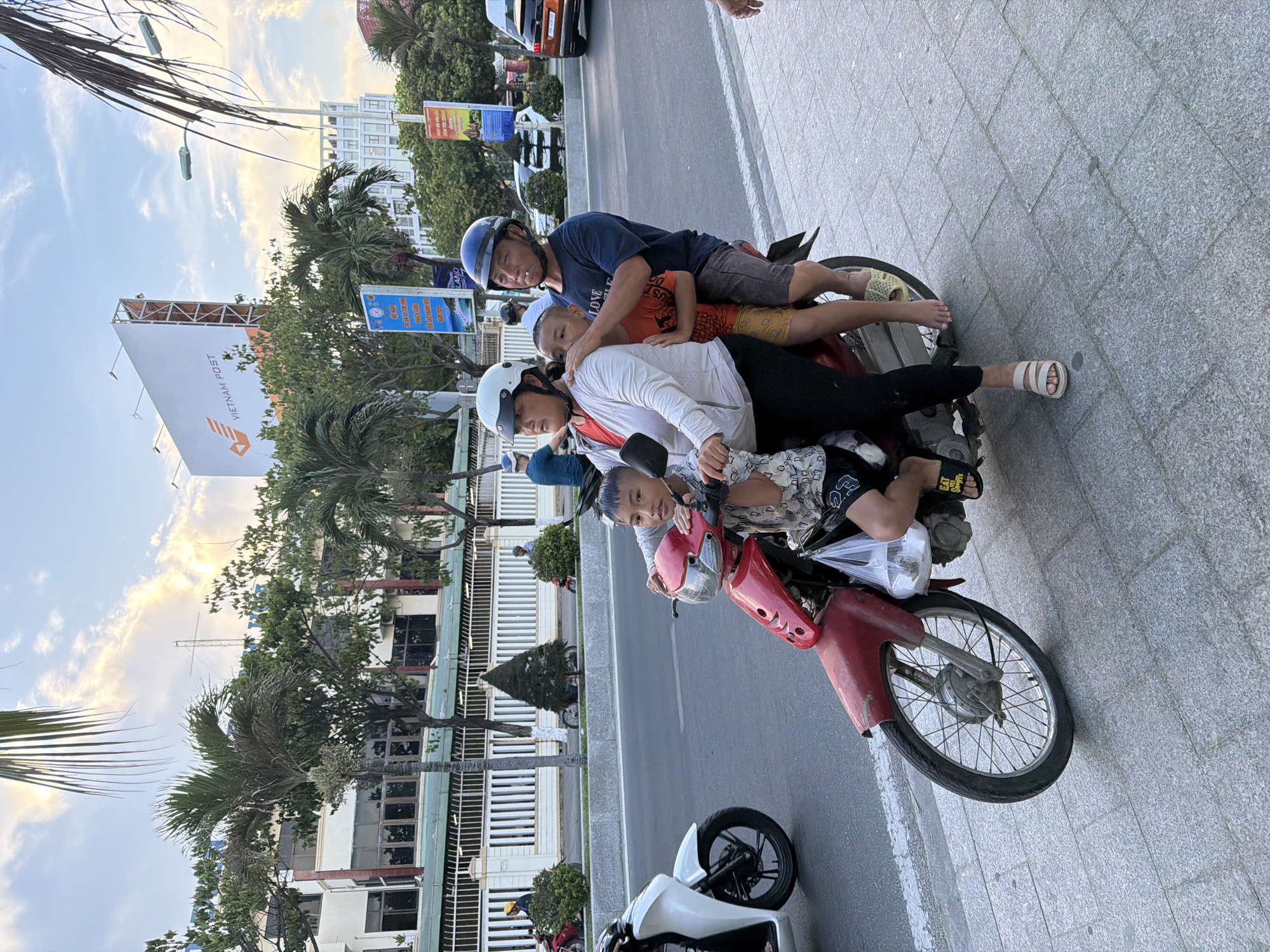 A family of four is riding on a red motorcycle on a city street in Nha Trang Ward, Vietnam. The father is driving, wearing a dark blue shirt and a blue helmet. The mother is in the middle, wearing a white jacket and a white helmet. Two young boys are seated between them. The older boy is wearing an orange shirt and yellow shorts, and the younger boy is wearing a patterned shirt and black shorts. The motorcycle is red with silver accents. It is daytime, and the sky is partly cloudy with bright sunlight. In the background, there are buildings, palm trees, and streetlights. A white motorcycle is parked on the left side of the image, and a white car is visible further down the street on the right. The motorcycle is on the road, and the family appears to be in motion.