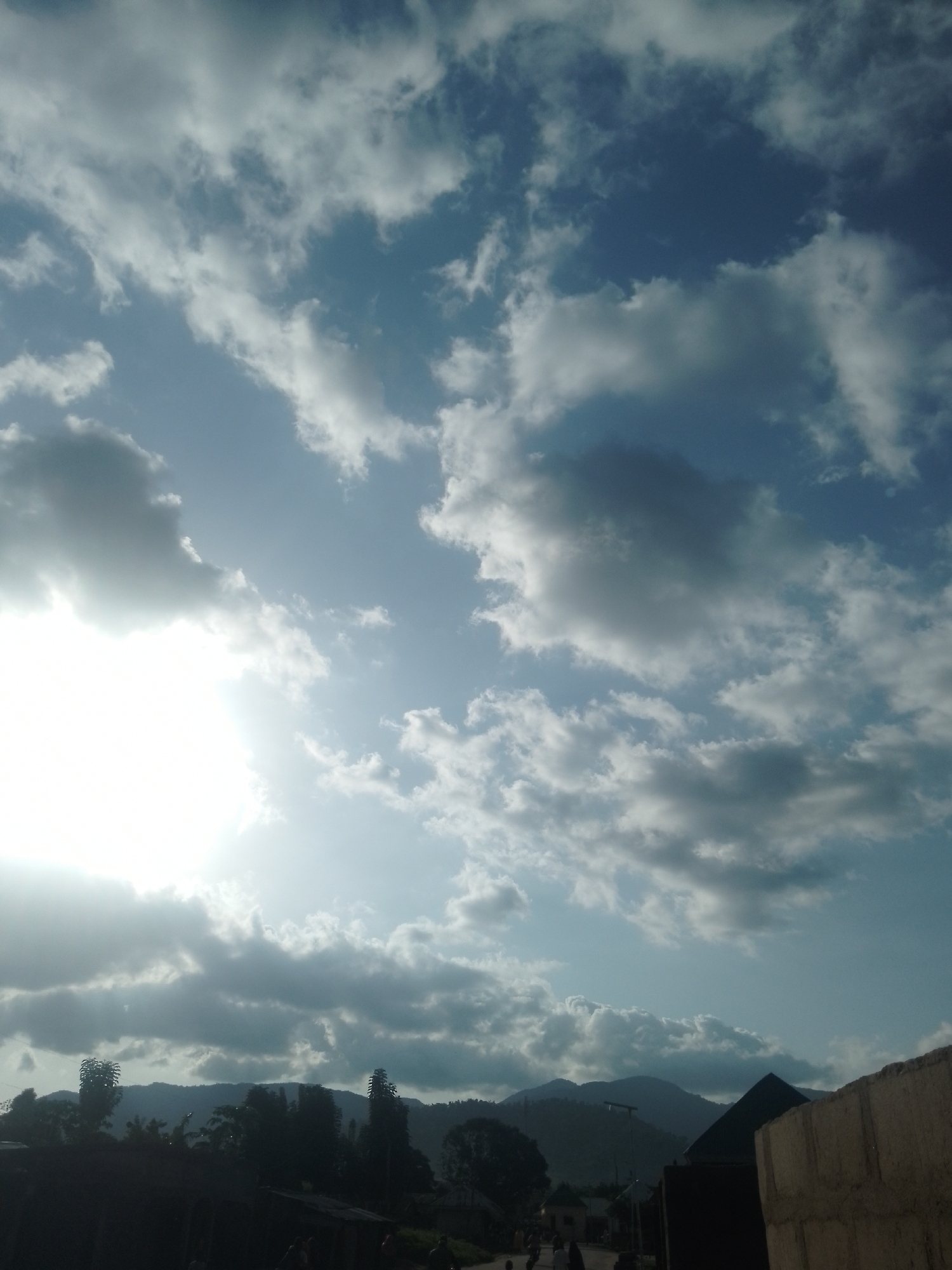 An outdoor vertical shot features a view of a mountainous landscape under a dramatic sky. The sky is a mix of deep blue and white clouds, with the sun partially obscured by clouds on the left side of the frame, casting a bright light. In the foreground, silhouetted against the sky, are the tops of trees and several buildings, some with dark roofs and walls. The overall impression is of a natural setting with signs of human habitation, possibly in a rural or suburban area. The location is described as Unknown, Unknown.