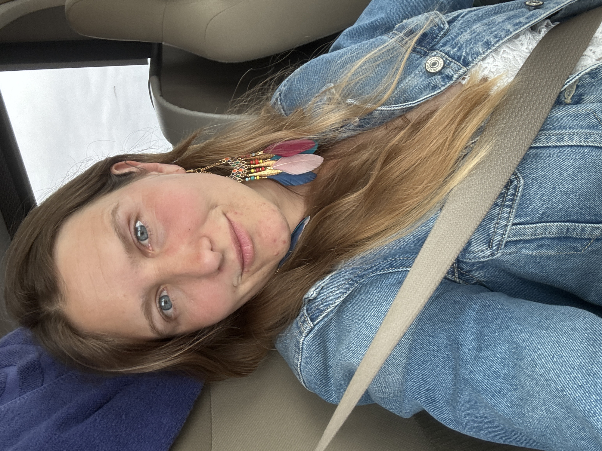 A woman is sitting in a vehicle, secured by a seatbelt. She is wearing a denim jacket over a white lacy top and colorful feather earrings. Her head rests on a purple fabric, possibly a towel or blanket. The car interior is visible with beige upholstery and a window partially visible on the upper left. The location is Unknown, Unknown.