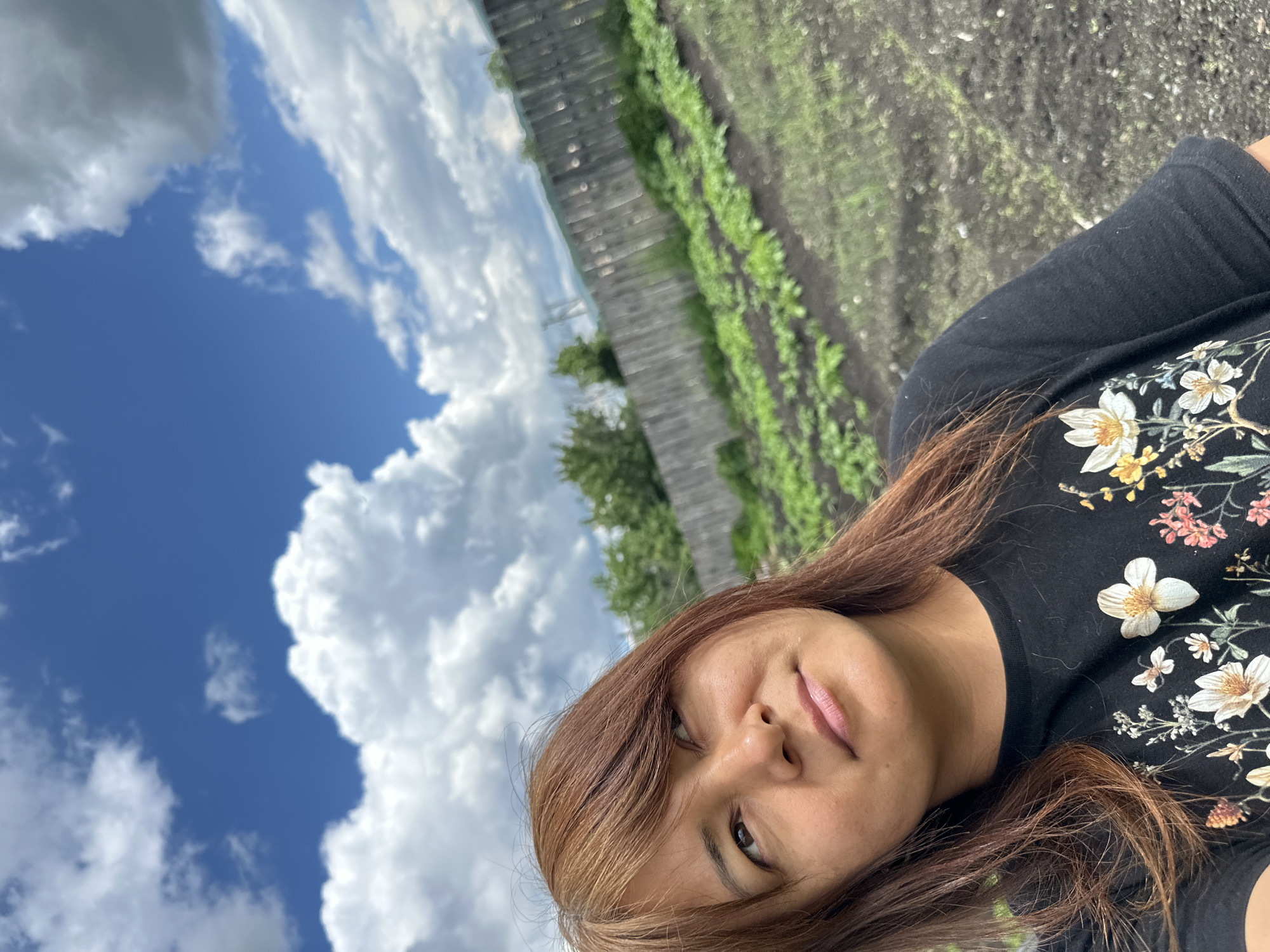 A medium shot selfie captures a woman with shoulder-length brown hair in a black t-shirt adorned with a floral pattern of white and yellow flowers and some pink and orange accents. She is looking upwards and to her right. The background consists of a garden with rows of green plants, a weathered wooden fence, and a sky filled with white, puffy clouds against a bright blue expanse. The scene is outdoors, likely in a rural or suburban setting, given the presence of a garden and a wooden fence. The lighting suggests it is daytime, with the sun providing bright illumination.