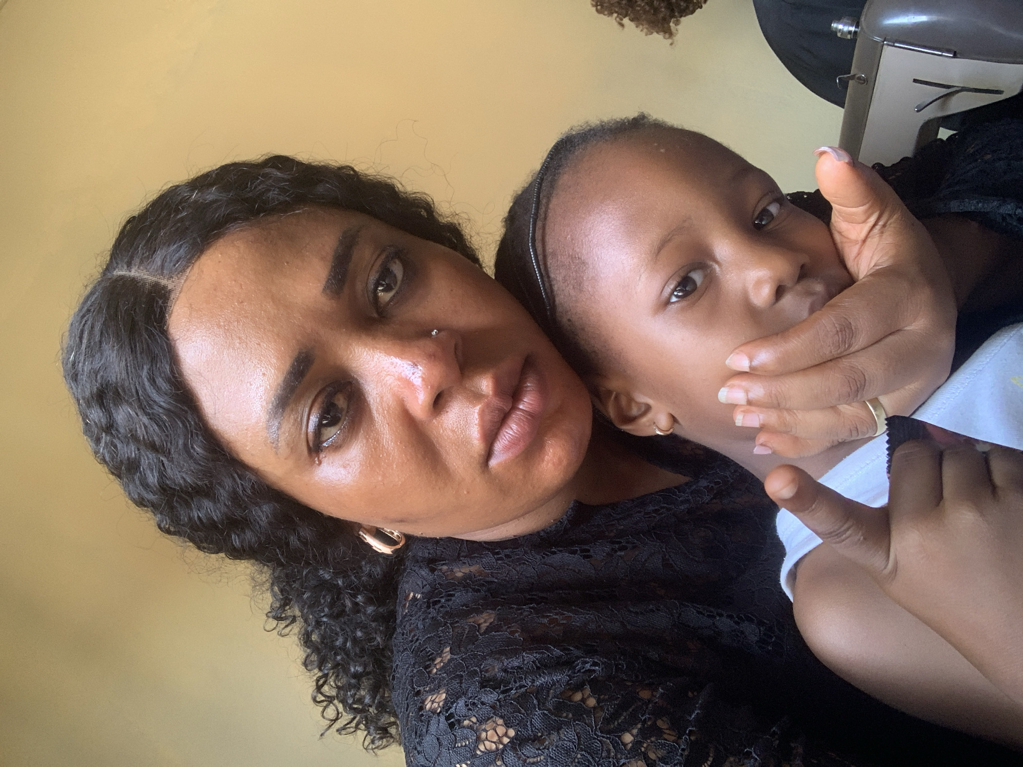 In an indoor setting in Sabon Tasha, Nigeria, a selfie-style photograph depicts a woman and a young child. The woman, with dark curly hair and a nose piercing, wears a black lace top and gold hoop earrings. She has a neutral expression. The child, with short hair held back by a headband, is held close, their face partially obscured by the woman's hand. The child wears a white garment. The background is a plain light yellow wall, with a glimpse of a sewing machine and other objects.