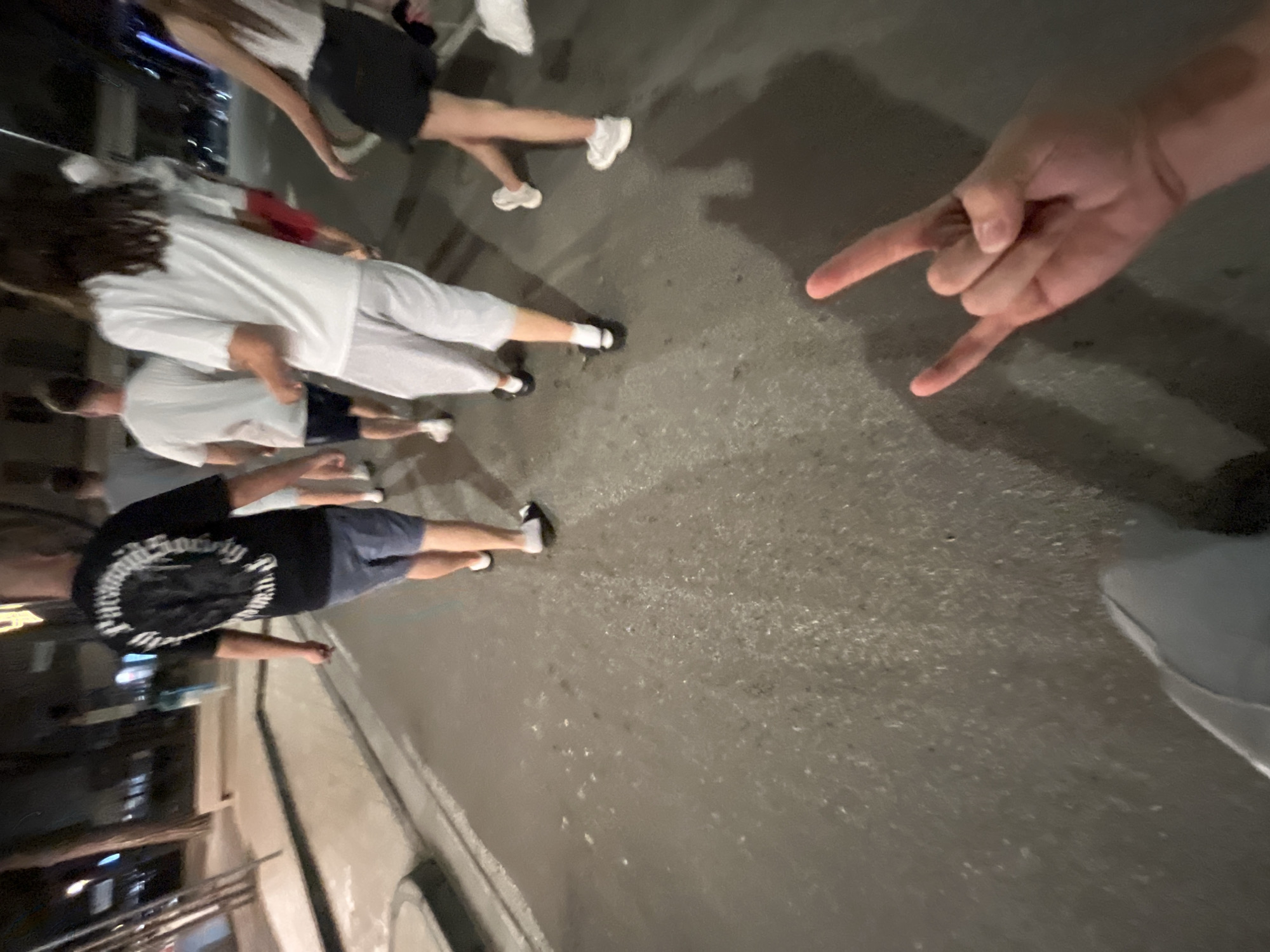 A ground-level, angled shot shows a group of at least six people walking on a paved street in Jesolo, Italy, with a hand making the "rock on" gesture in the foreground.
The people appear to be casually dressed. One person wears a black t-shirt with white writing on the back. Another wears a white shirt and light-colored pants. A third person wears a light-colored shirt and dark shorts. They are walking parallel to the street edge, near a building.
The hand making the "rock on" gesture is in the right side of the frame. A shoe appears near the bottom right of the frame.
The environment is dimly lit, suggesting it is nighttime. A building is on the left side of the frame, with some lighting and architectural details visible. The road takes up the middle of the frame.