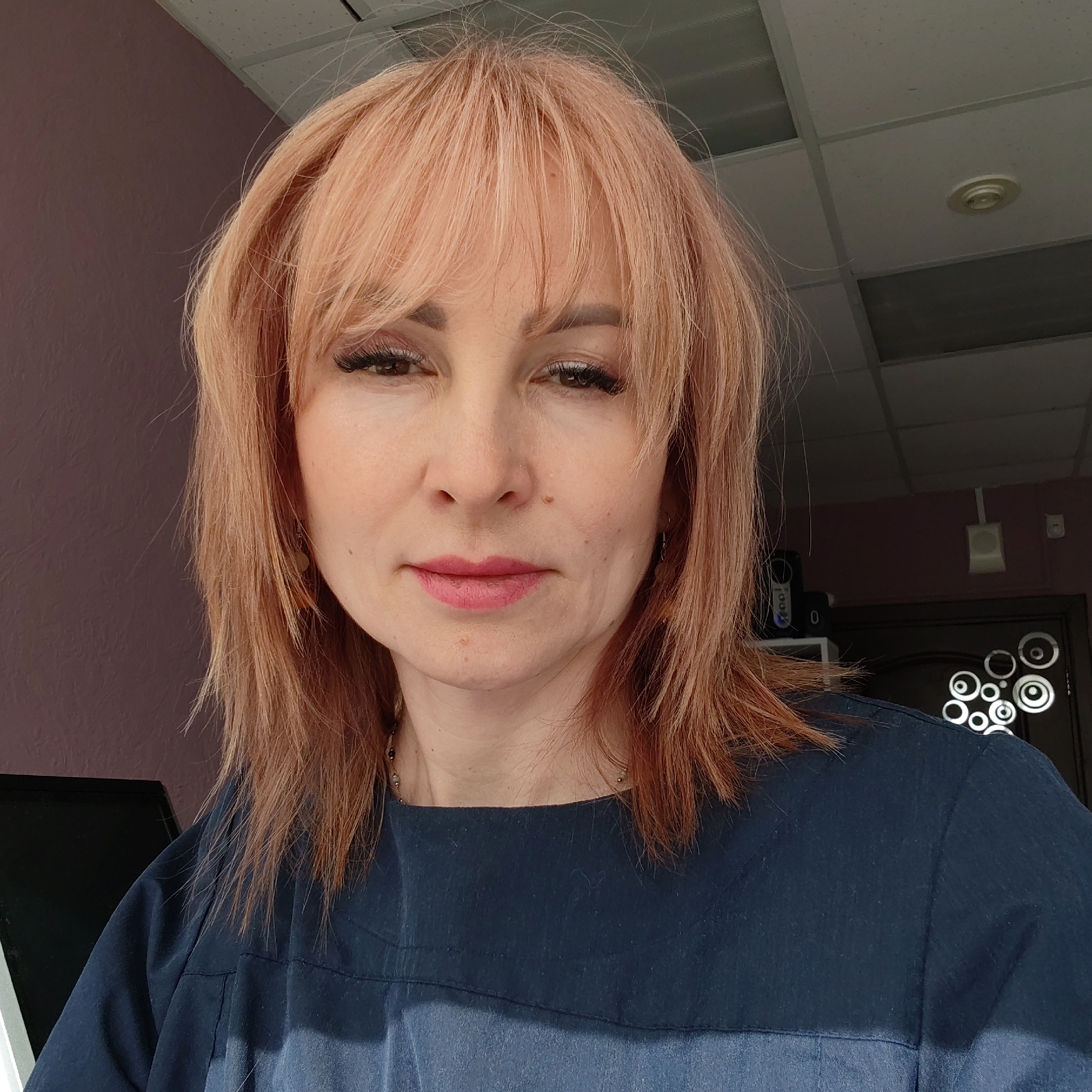A middle-aged woman is centered in the frame, with a close-up perspective focusing primarily on her upper torso and face. She has light pink hair with bangs and is wearing a dark blue top. Her makeup is noticeable, with defined eyelashes and pink lipstick. Her facial expression is neutral.
The background reveals interior details. A portion of a pink-colored wall is visible to the left of her. To the right, there is a door with a geometric design, partially obscured by a shelf-like structure that holds a speaker. Above her, the ceiling is comprised of white tiles.
The image was taken indoors, possibly in the city of Chaikovsky, Russia.