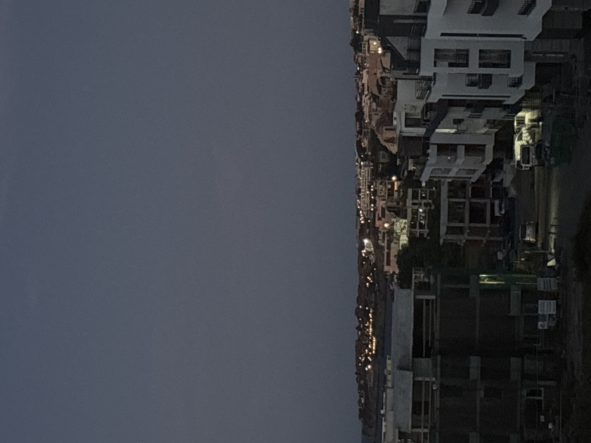 The image depicts an elevated view of a coastal town at dusk or dawn. The sky is a deep, muted blue-gray, suggesting low light conditions. In the foreground, the silhouette of a dark building under construction is visible on the left, with exposed scaffolding and concrete structures. To the right, a row of modern buildings, illuminated by interior lights, line a street. These buildings feature balconies and large windows. Further into the distance, a more densely populated area of buildings with illuminated windows stretches along the coastline. The overall impression is one of a developing urban area by the sea during twilight.