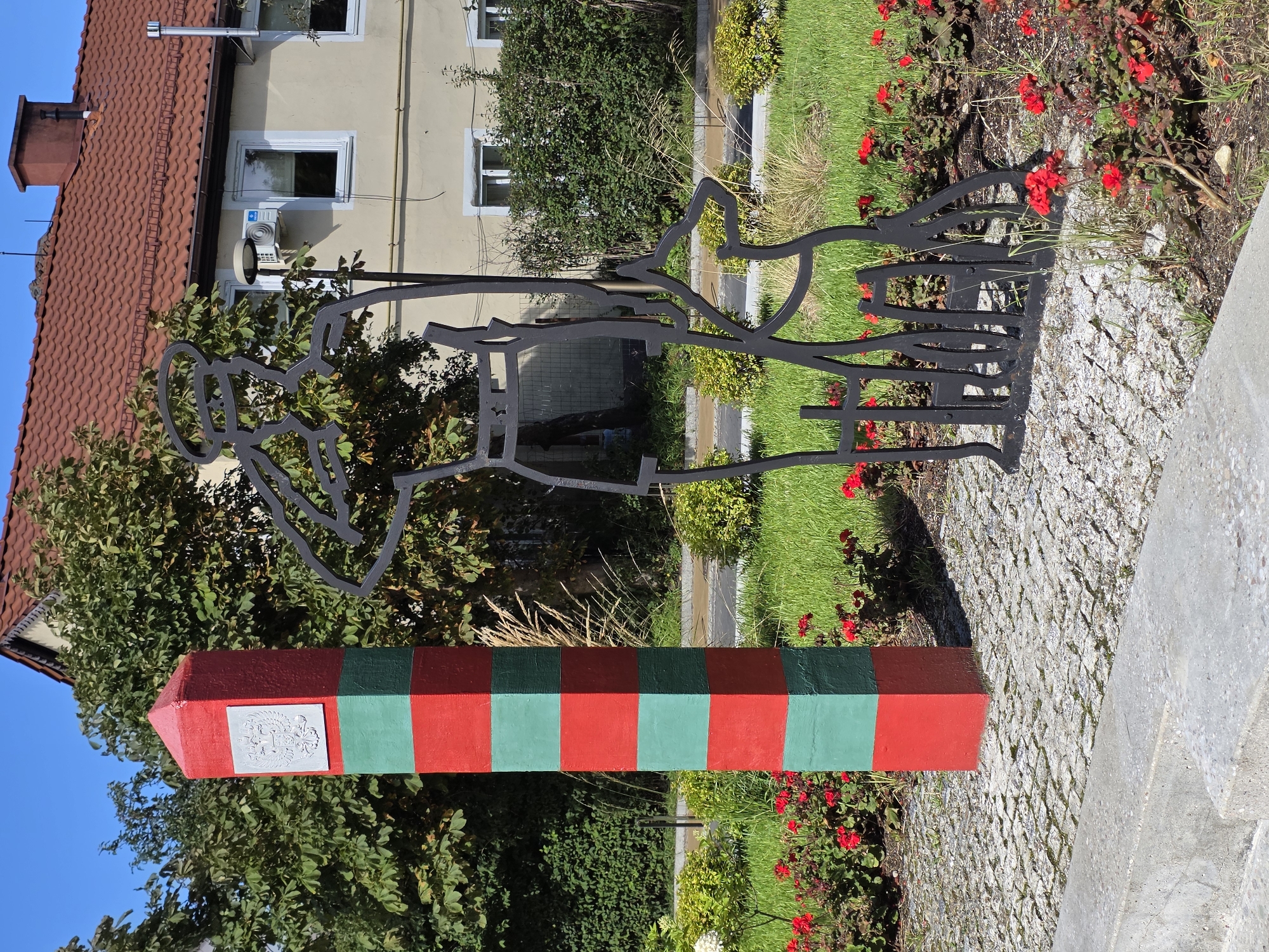In Zelenogradsk, Russia, on a paved area next to a sidewalk, stands a red and green striped border post. A silver emblem is affixed to the top of the post. To the right of the post, a black metal silhouette depicts a person, possibly a border guard with a dog, standing with their back to the viewer. The silhouette is positioned in a grassy area with red flowers and small green bushes. In the background, a light-colored building with a red tiled roof and white-trimmed windows is visible. The sky is clear and blue.