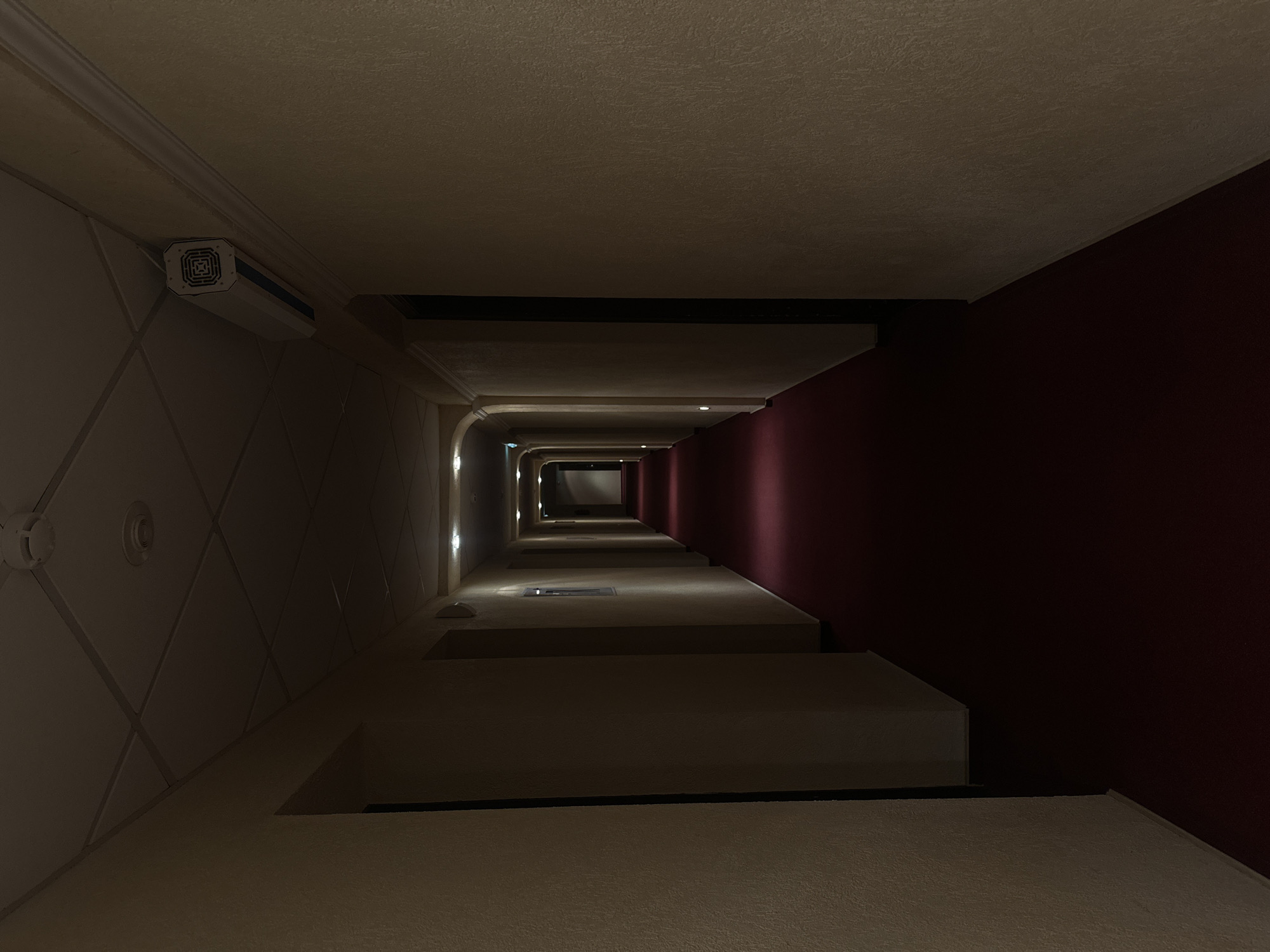 The image depicts a long, straight hallway in Perm, Russia. The hallway appears to be in a building, possibly a hotel or office complex. The walls are beige with a darker burgundy section on one side. The ceiling is low and evenly lit. The hallway recedes into the distance, with visible doorways and lights illuminating the path. The angle of the shot suggests it was taken from a high vantage point, looking down the hallway. There are ventilation systems mounted to the walls.