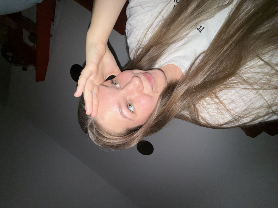 A woman with long, light brown hair and green eyes poses for the camera. Her hand is near her forehead. She wears a white shirt with some text. The background is dark, showing a ceiling with two black circular objects attached. Part of a red object can be seen in the background as well. The image has been rotated.