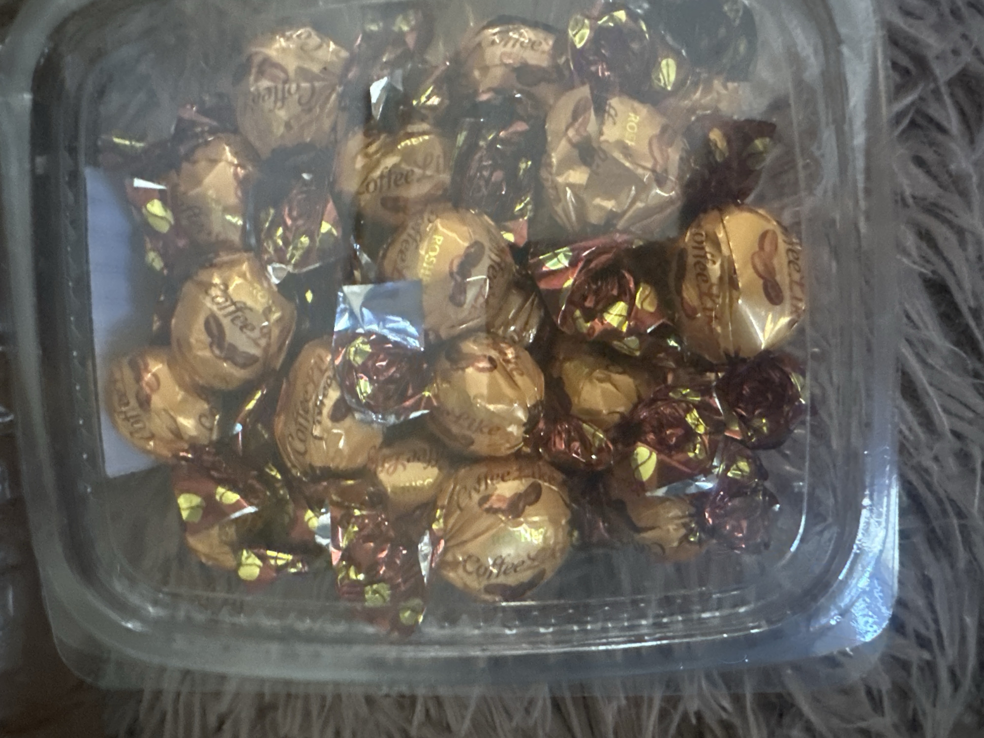 An overhead, slightly blurry, close-up shot shows a clear plastic container filled with individually wrapped candies. The candies are predominantly gold and maroon foil-wrapped, with some displaying the text "Coffee" and what appears to be coffee beans. The container is resting on a soft, fuzzy, light-colored surface, possibly a rug or blanket. The background is out of focus, but appears to be a dark, indistinct surface. The lighting is diffused, casting a soft glow on the candies. There are no people visible in the image. The scene is indoors, likely a home environment.