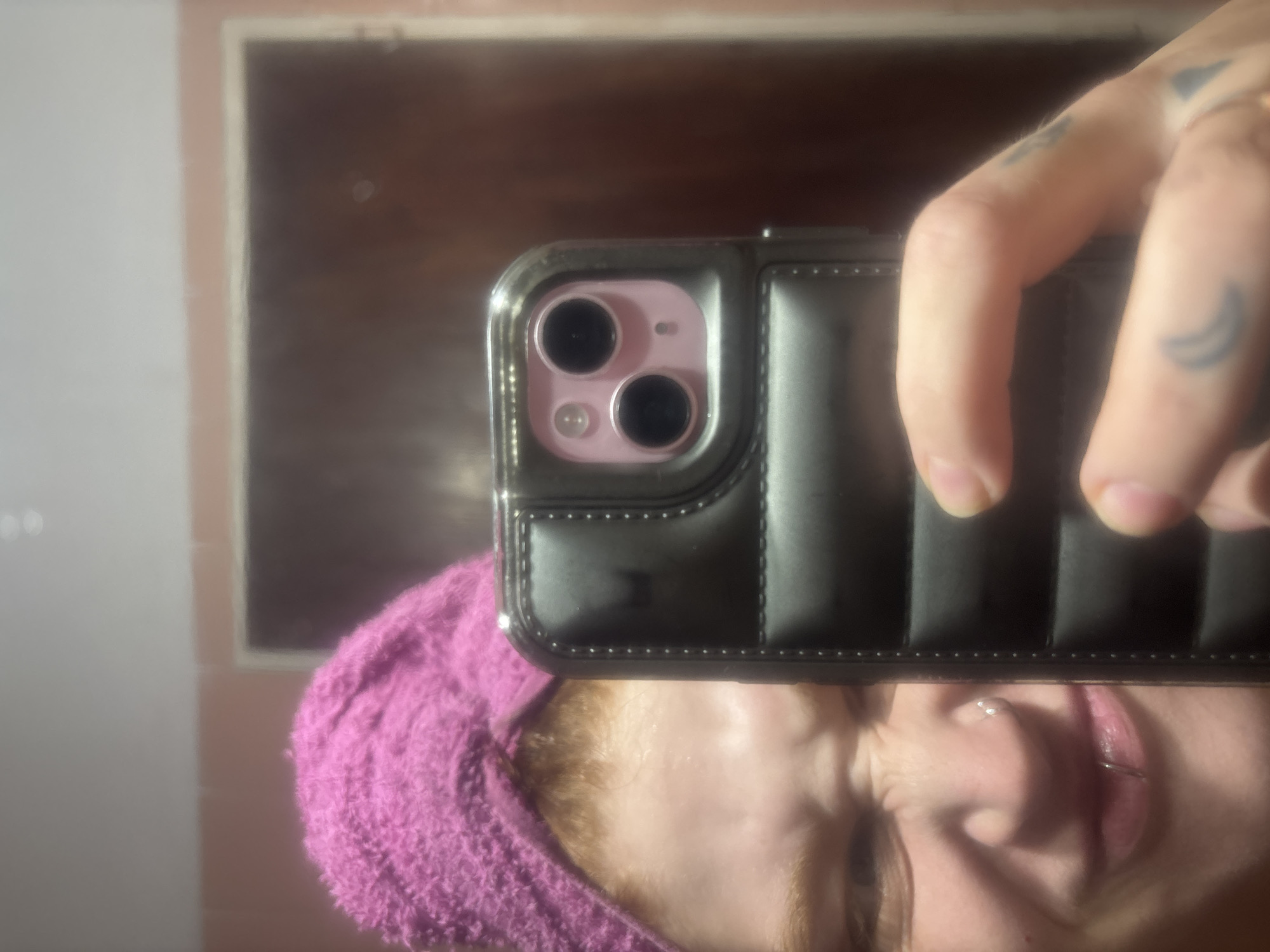 A close-up selfie taken in a bathroom setting, likely indoors due to the artificial lighting. The subject is holding a black smartphone, an iPhone 13 with a pink camera housing, obstructing part of their face. The person has fair skin, blonde hair covered by a purple towel, and visible piercings on their nose and lip. A hand with multiple tattoos is holding the phone. The background features a mirror and light-colored walls, with a blurry reflection of a window or an object. The exact location is difficult to determine.