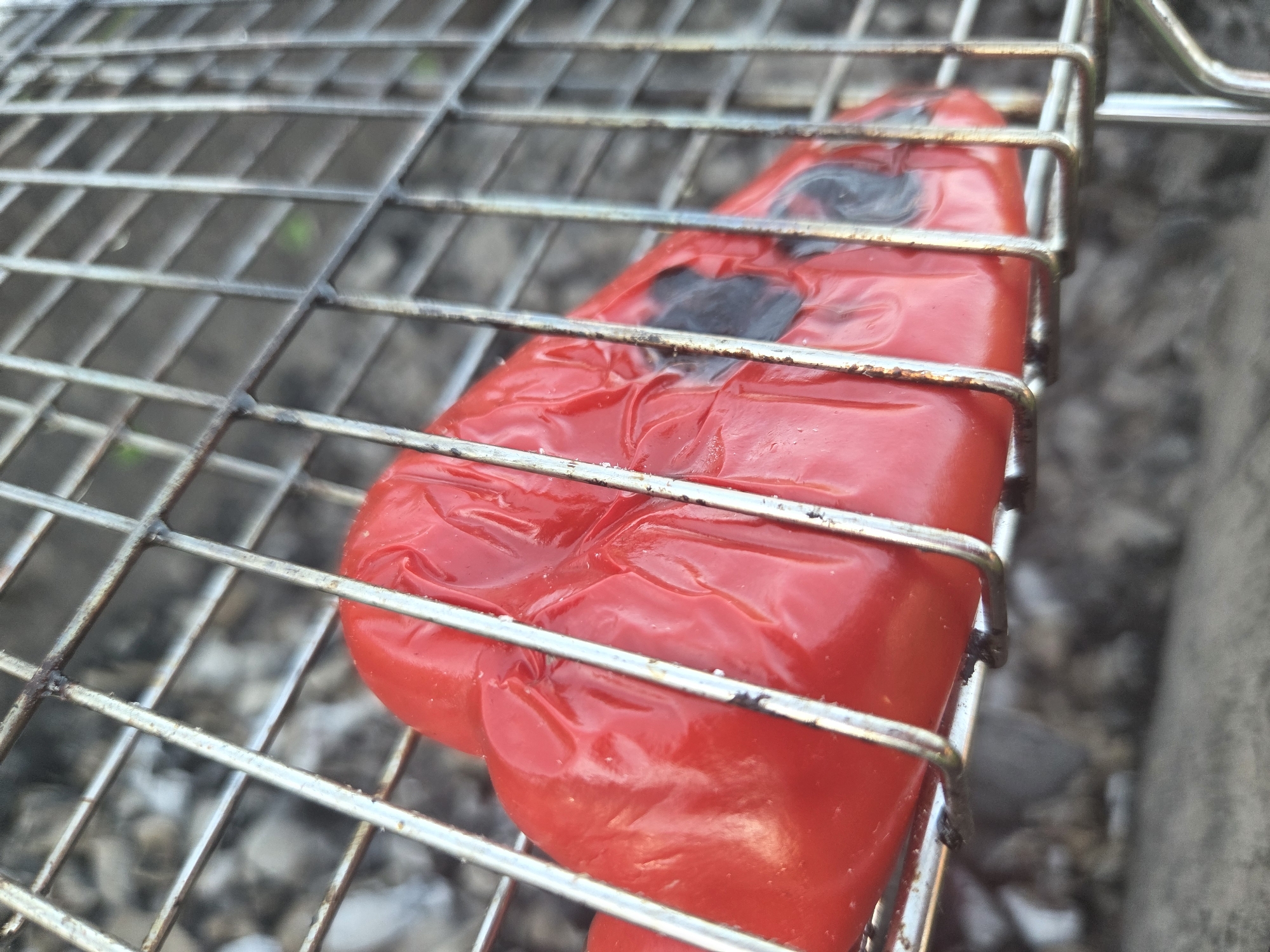 A close-up, angled shot shows a red bell pepper on a metal grill. The pepper appears to be cooking, with some charred spots visible. The grill is made of thin metal bars and is positioned over what looks like an outdoor surface, possibly charcoal or dirt. The background is blurred, but suggests an outdoor environment. There are no people visible in the image. The scene is likely in Frolovo, Russia.