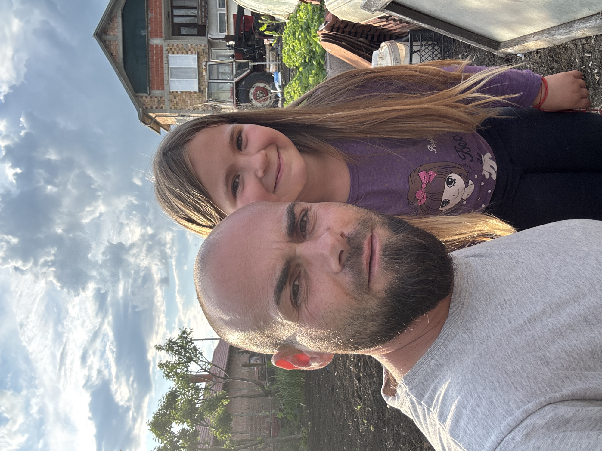 A man and a girl are posing for a selfie. The man has a shaved head and a beard. The girl is standing behind him and has long brown hair. She is wearing a purple shirt. In the background is a house made of red and white bricks, farm equipment, and foliage. The location may be Sarnevo, Bulgaria.