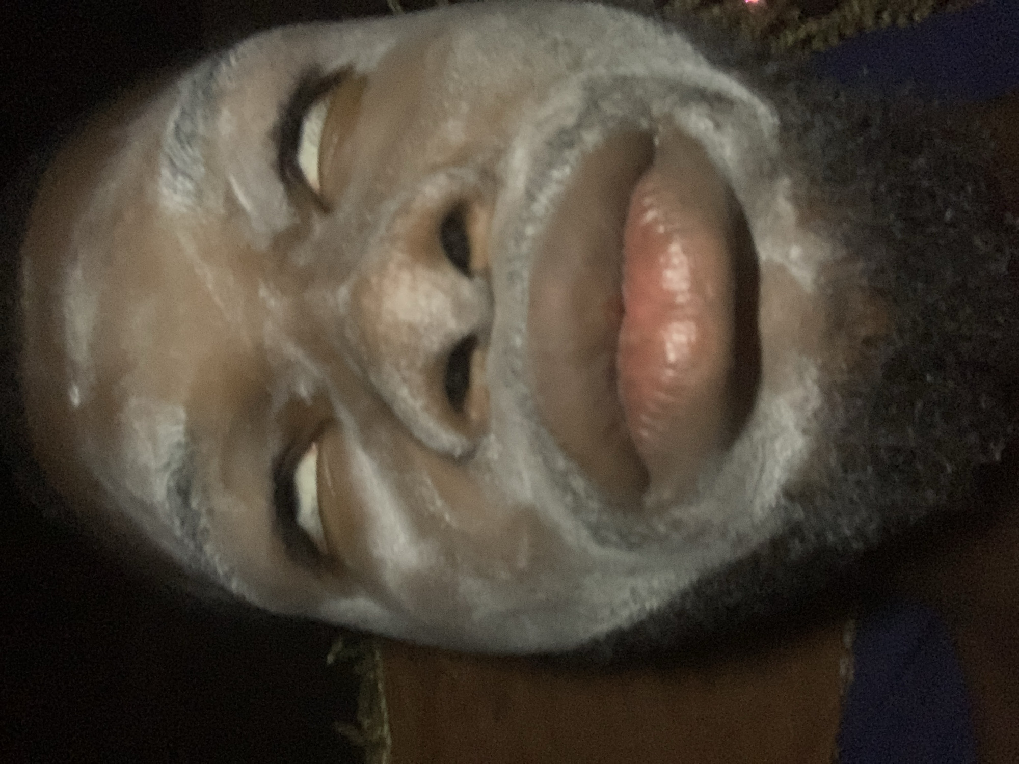 A man's face fills the frame. His eyes are open but unfocused, and his mouth is open wide, revealing his tongue. A white substance covers much of his face, resembling a facial mask. He has a dark beard. The image is poorly lit, casting dark shadows. Location is not discernible from the image.