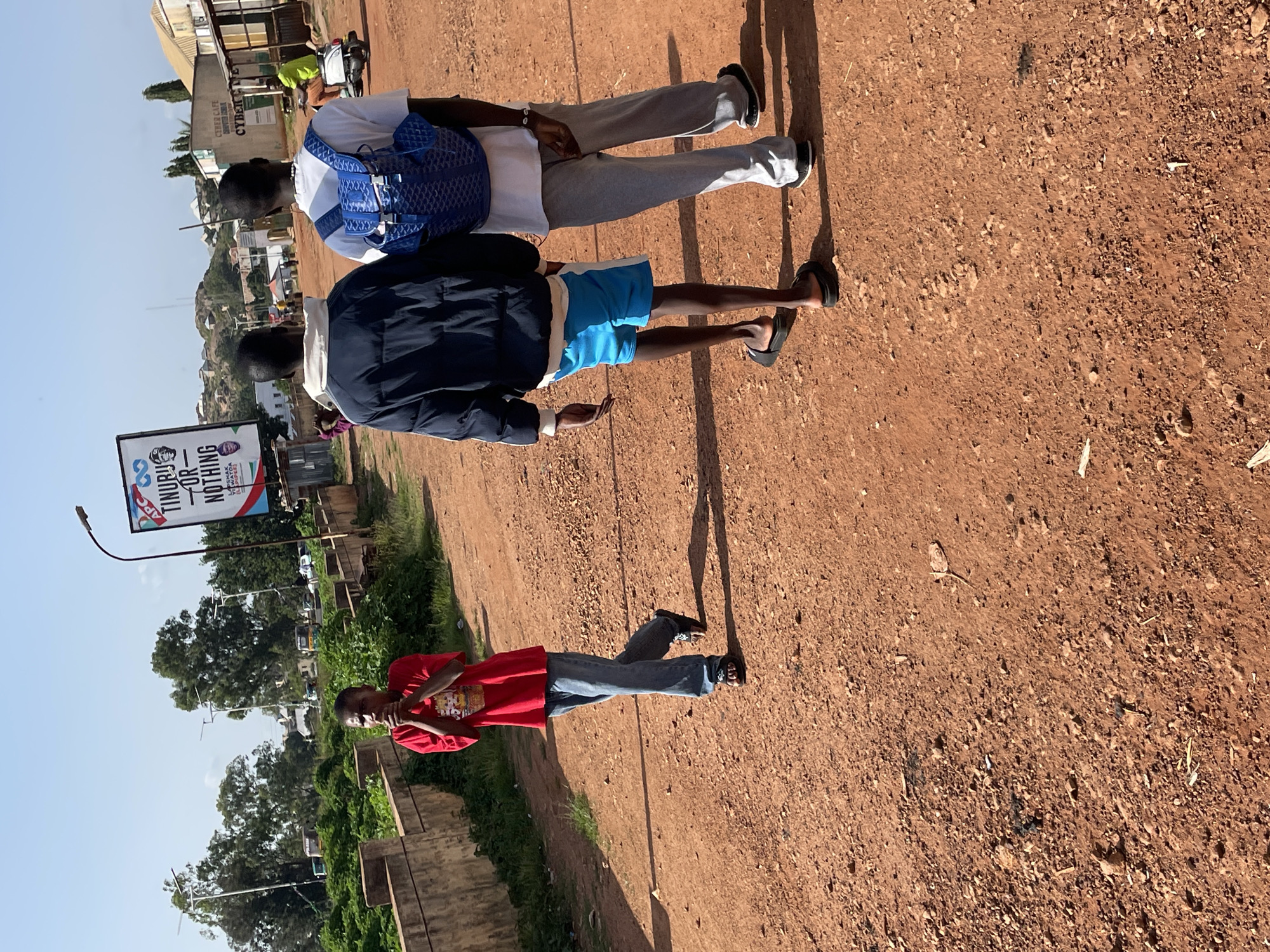 In Gura Top, Nigeria, on a dirt road, three young men are walking. The first, on the left, wears a red t-shirt and blue jeans. The second, in the middle, is dressed in a dark jacket and blue shorts. The third, on the right, sports a white t-shirt and gray sweatpants, carrying a blue backpack. The background features trees, buildings, and a billboard with text and images. A person is also visible riding a scooter in the distance. The ground is covered in reddish-brown dirt with scattered rocks and debris. The sky is clear and blue.