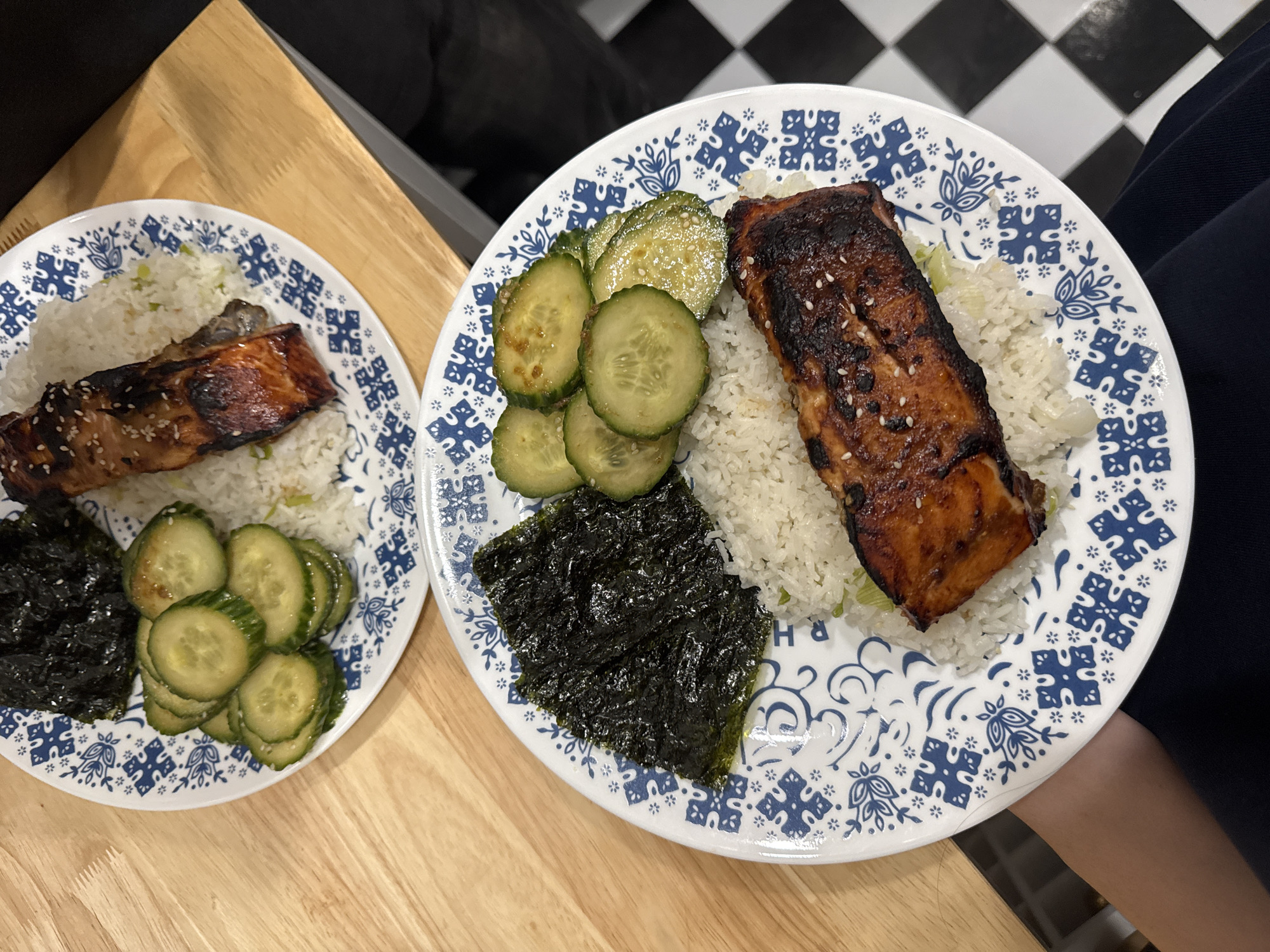 The image shows two plates of food, likely home-cooked. Each plate has a serving of white rice topped with a piece of grilled salmon. Alongside the rice and salmon, there are cucumber slices and a dark, crumbled seaweed salad. The plates are decorated with a blue and white floral pattern. One plate sits on a light wooden surface, while the other is held by a person wearing a dark blue sleeve. The background includes a black and white checkered floor, possibly indicating a kitchen setting. The location in the city of New York, United States of America cannot be determined from the image.