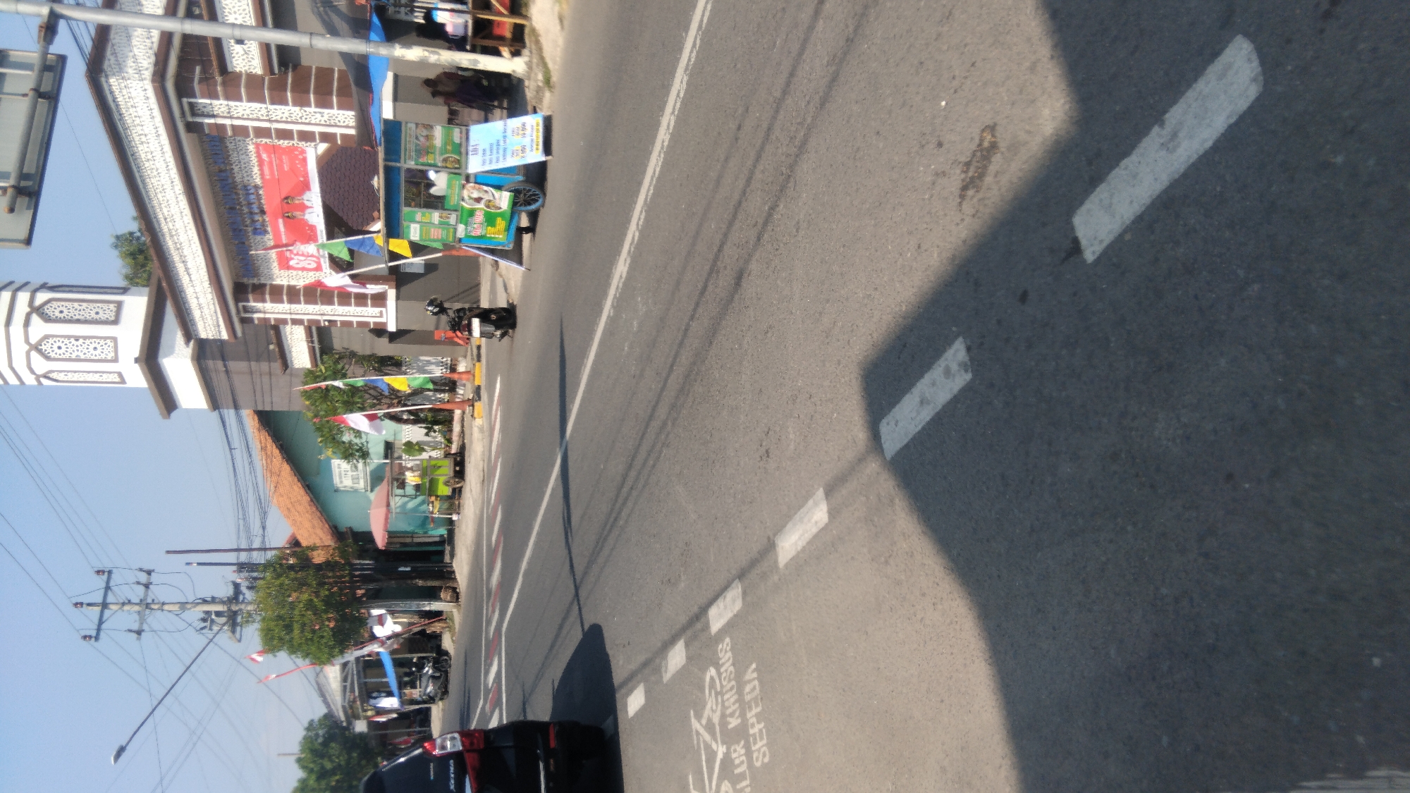The image depicts a street scene in Proyonanggan Tengah, Indonesia. The foreground shows a paved road with white dashed lines and a designated bicycle lane marked with the words "JALUR KHUSUS SEPEDA" and a bicycle symbol. A dark-colored SUV is partially visible on the left side of the road, facing away from the camera.
In the mid-ground, a sidewalk runs alongside the road, with several small food stalls and vendors. One stall has a blue and green cart with various items displayed, and a sign with text and prices. Indonesian flags are visible, attached to poles and some stalls. Behind the stalls, there are buildings, including a mosque with a tall minaret and decorative white patterns. Another building to the left has a tiled orange roof.
The background shows a clear blue sky, electrical poles with wires, and trees. Several motorcycles are parked on the sidewalk or near the stalls. People are visible in the distance, some standing near the stalls. The overall scene suggests a typical day in a busy street with commercial activity, common in Indonesian towns.