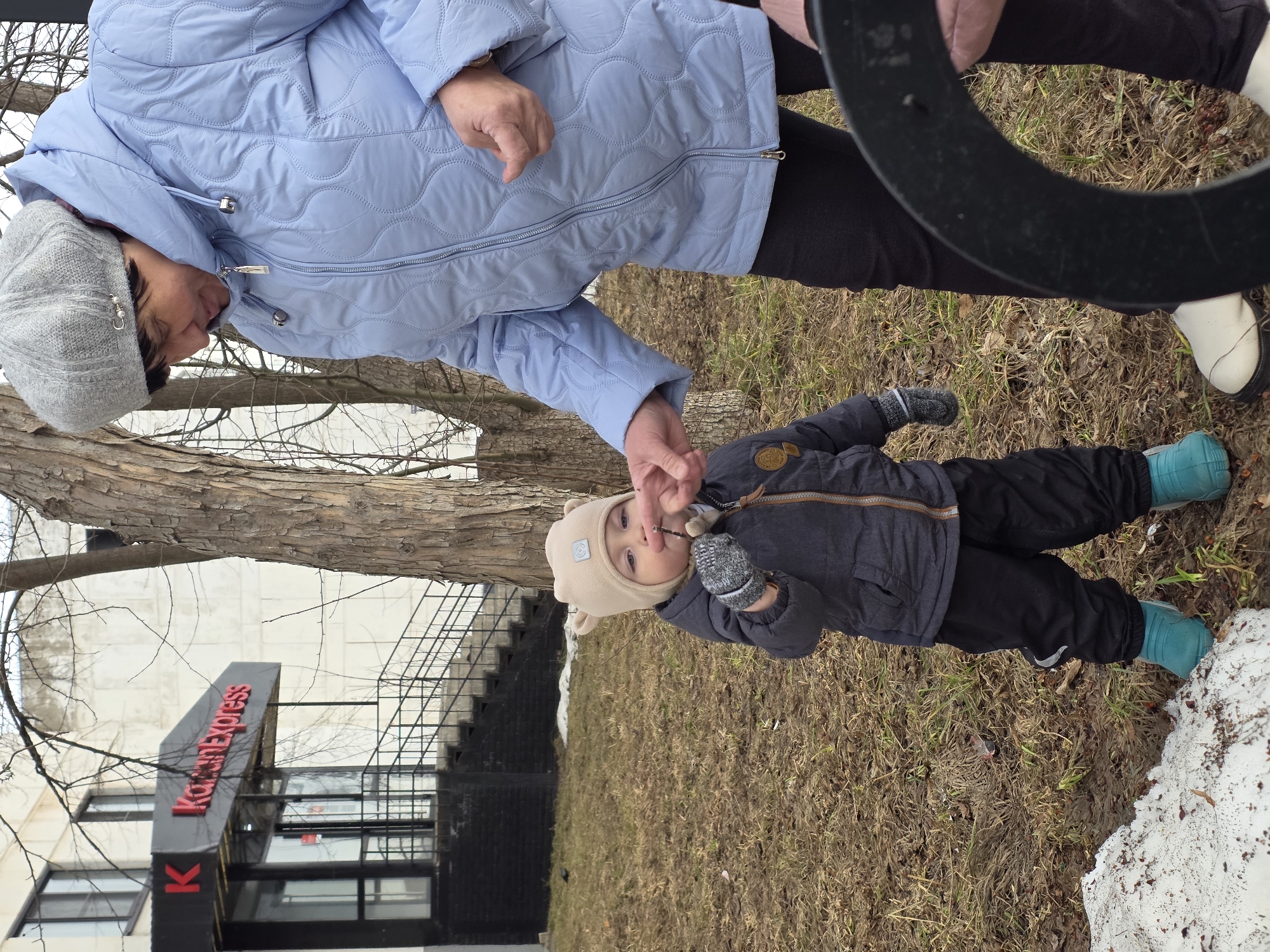 The image captures an outdoor scene in Chaikovsky, Russia, featuring an adult female and a young child. The adult, positioned on the right, wears a grey knitted beret, a light blue quilted puffer jacket, dark trousers, and white shoes with black soles. She is looking downwards, extending her right hand towards the child. The child, standing on the left, wears a beige hat with ear-like features, a dark grey puffer jacket, dark trousers, turquoise boots, and grey mittens. The child holds a small, dark, twig-like object between the thumb and forefinger of their right hand, examining it.
In the background, a light-colored building with dark-framed windows is visible. An entrance with black stairs and a black metal railing leads up to an awning displaying "KazanExpress" in red lettering, preceded by a red "K". The lower facade of the building around the entrance is dark brick. A large, bare tree trunk stands prominently between the individuals and the building. The ground is covered with dormant, dry brown grass and scattered patches of melting snow. A dark, curved object is partially visible in the lower right foreground, obscuring part of the adult's legs and feet. The scene suggests late winter or early spring conditions.