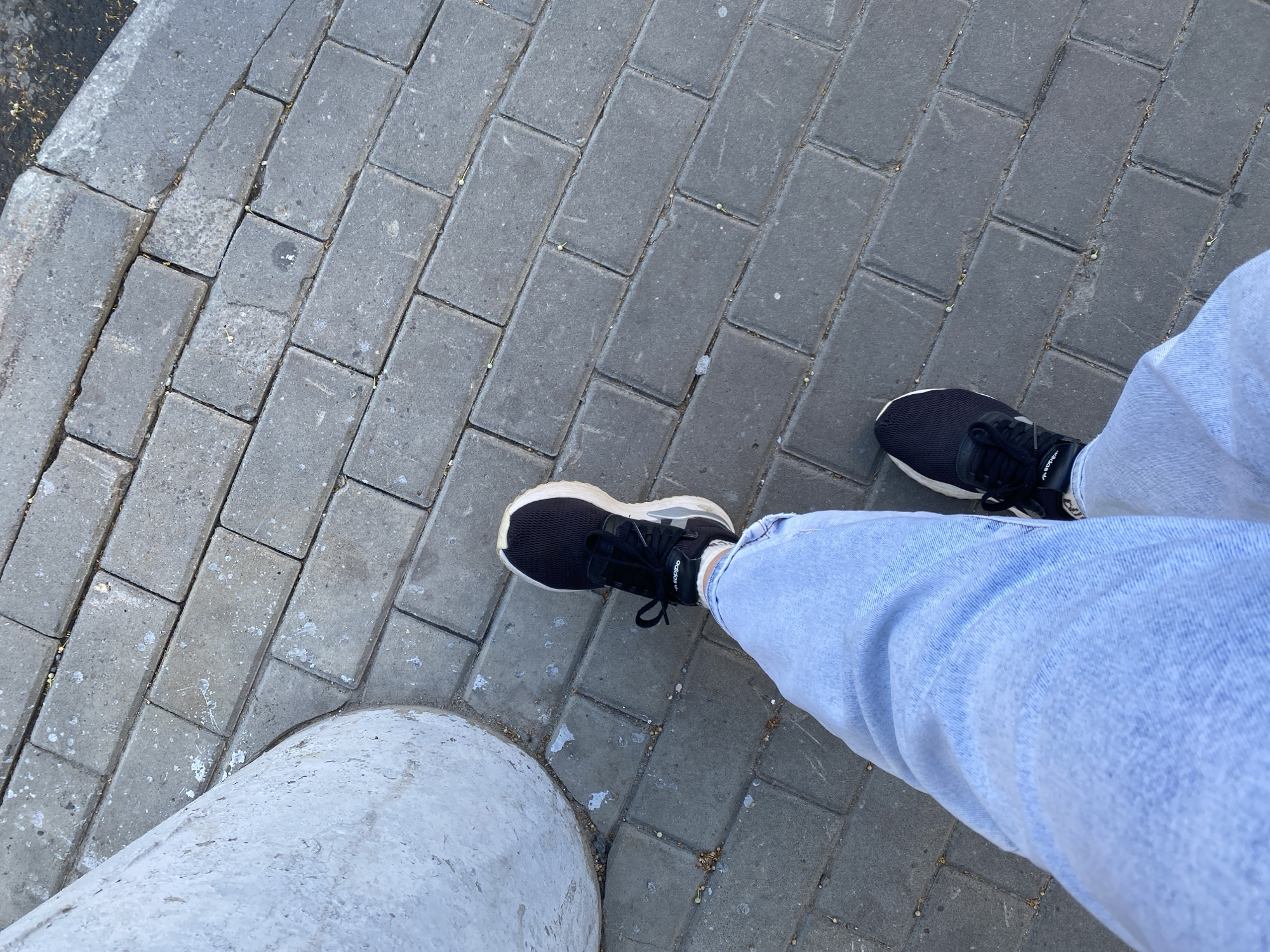 The image shows a person's legs and feet from a top-down perspective, standing on a gray paved surface composed of rectangular bricks. The person is wearing light blue jeans and black athletic shoes. The shoes have white soles and black laces, and the brand name "adidas" is visible on the side of one shoe.
The scene appears to be outdoors, on a sidewalk or walkway. To the left of the frame, a portion of a rounded, light-colored concrete structure, possibly a bollard or a part of a planter, is visible. The paving stones are somewhat worn and show some scattered debris.
The time of day is not explicitly indicated, but the lighting suggests it is daytime, possibly morning or afternoon, with diffused sunlight. The weather appears to be dry. The location is identified as Novaya Usman, Russia. No specific emotional cues are conveyed by the image, as only the lower half of the person is visible. The overall impression is of someone standing on a street or pathway.