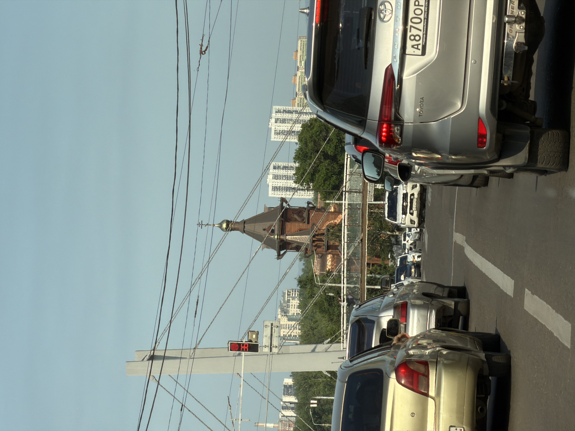The image shows a traffic jam on a sunny day in Krasnoyarsk, Russia. Several cars are lined up on a road, with a golden-colored car in the foreground on the left and a silver Toyota SUV on the right. In the background, there are multi-story apartment buildings and a wooden church with a golden dome. A pedestrian bridge spans across the road in the middle distance. Traffic lights are visible, indicating a stop or slow down. The sky is clear and blue. The environment appears to be urban, with evidence of infrastructure like power lines and traffic signals.