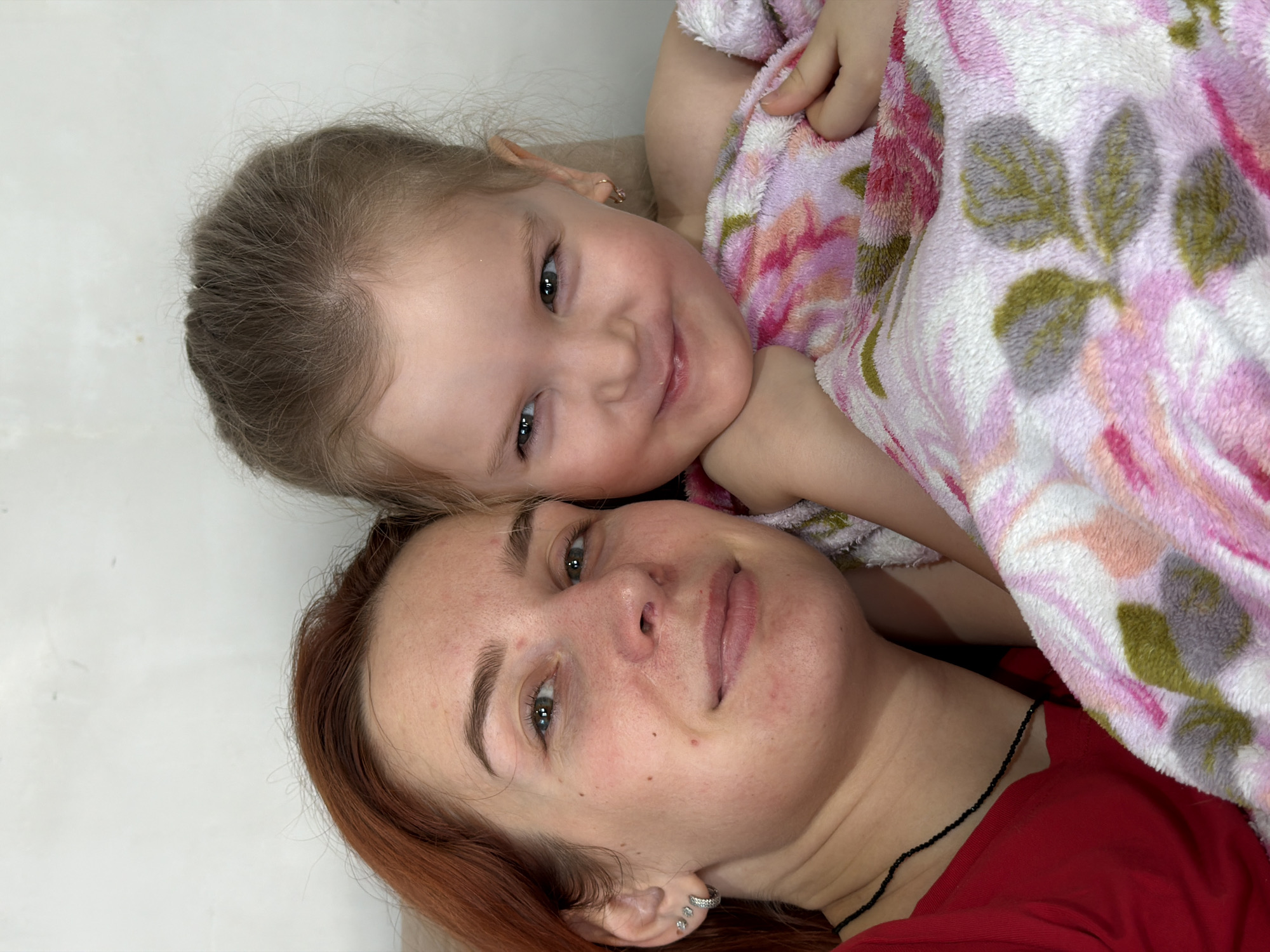 This close-up portrait, originating from Chaikovsky, Russia, features two individuals: an adult female and a young female child. The adult, positioned on the left, has reddish-brown hair and greenish-blue eyes, looking directly at the camera with a slight smile. She wears a red garment, a thin black beaded necklace, and three silver-colored stud earrings in her left ear. Her face shows minor blemishes/freckles. The child, situated on the right, has light brown hair and blue eyes, looking slightly to the viewer's left with a wide, playful smile. She has a small gold-colored hoop earring in her right ear. The child is partially wrapped in a soft, fuzzy blanket featuring a floral pattern in pink, white, green, and gray hues, depicting roses and leaves. Both individuals are positioned in close proximity, with the adult's head tilted towards the child. The background consists of a plain, light-colored wall with minor surface imperfections. No other objects or environmental details are discernible.