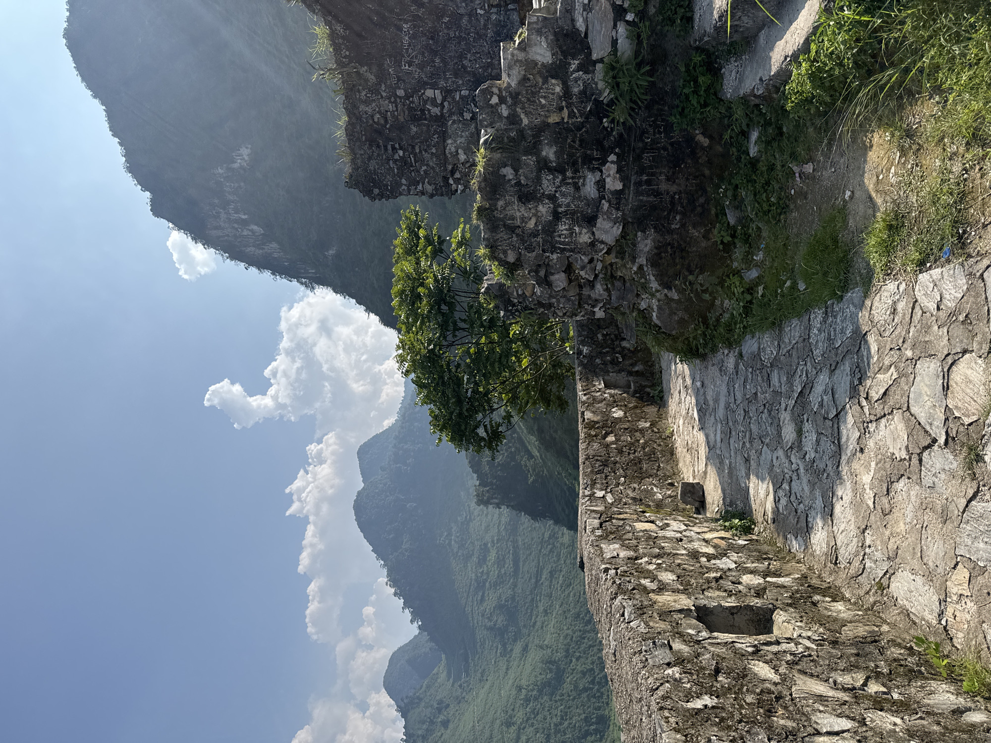 The image shows a stone walkway and walls overlooking a mountainous landscape in Đường Thượng Commune, Vietnam. The walkway is constructed of irregular stones and runs along a stone wall. Beyond the wall, a steep, forested mountain range rises into a blue sky with scattered clouds. A small tree grows near the wall, partially obscuring the view of the mountains. The scene is bathed in sunlight.