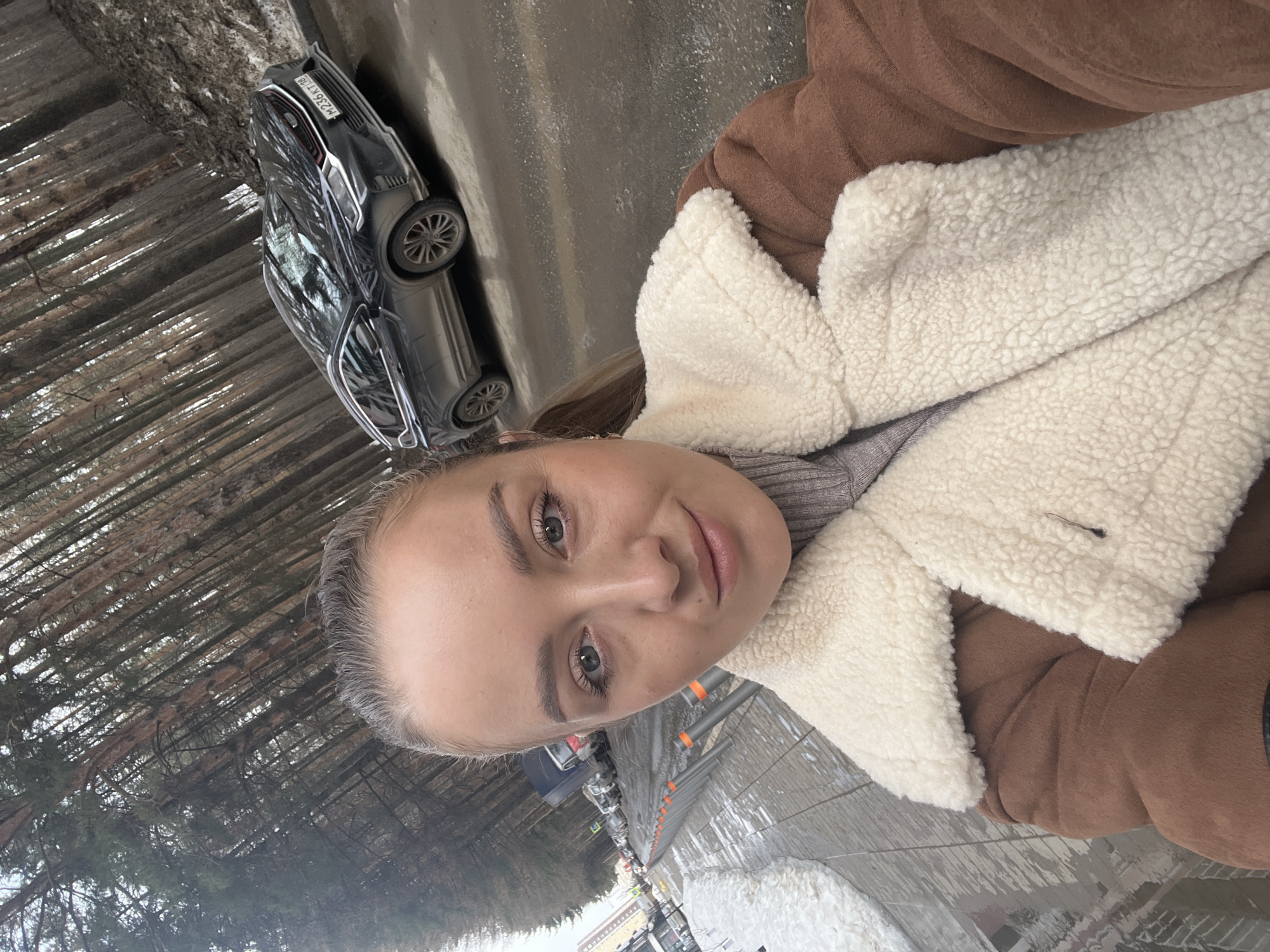 A female subject is depicted in a close-up selfie outdoors, positioned on an urban street bordering a coniferous forest in Unknown, Unknown. The subject, with fair skin, light-colored eyes, and light brown hair pulled back, is wearing a brown suede or faux-suede jacket with a cream-colored sherpa lining and collar over a light grey ribbed knit top. She is facing the camera, exhibiting a slight smile.
Behind and to the right of the subject, a dark grey Geely crossover SUV (model identification consistent with Coolray/Atlas Pro) is parked on the shoulder of the road, partially on a dirt and melting snow bank. The vehicle's Russian license plate, "M236KT 18", is clearly visible on the front. The road surface is dark asphalt, appearing wet, and partially covered with dirty, melting snow. To the left, a paved sidewalk, likely constructed from concrete pavers, runs parallel to the road. This sidewalk is bordered by a series of silver bollards featuring orange tops. Patches of dirty, melting snow are present on the sidewalk and along its edge. In the background, a dense stand of tall, slender coniferous trees forms a forest, with other parked vehicles, including a blue box truck, visible further down the street. The sky is overcast. The overall environment indicates late winter or early spring conditions with thawing snow.
