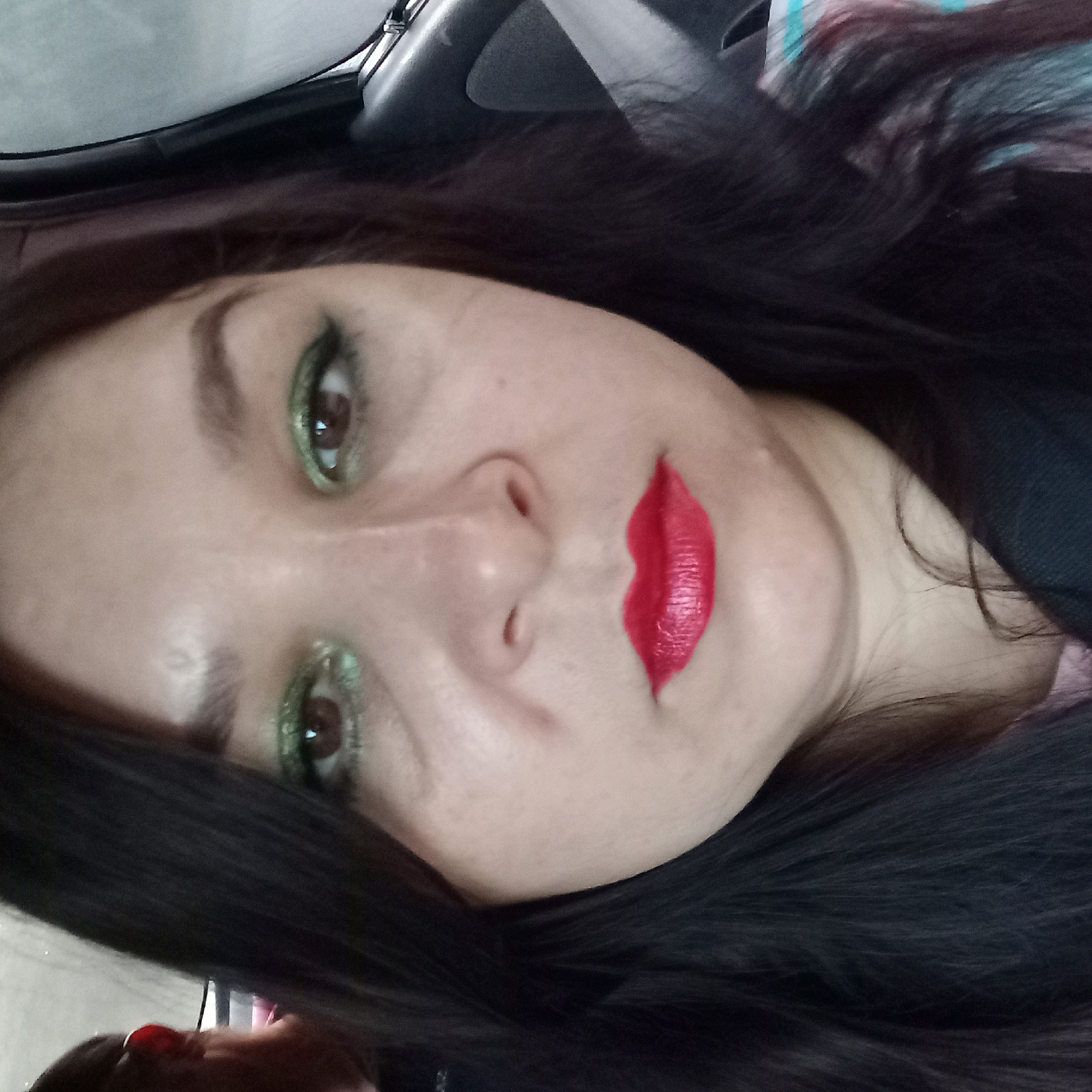 The image is a close-up selfie of a woman with dark hair, wearing green eyeshadow and bright red lipstick. She appears to be in a car, suggested by the presence of a seatbelt buckle and a car window frame. The background is blurry, showing a hint of the exterior environment. There is no indication of the specific location within Kropyvnytskyi, Ukraine, or any other geographical details. The woman is looking directly at the camera, with a neutral expression. The lighting suggests it is daytime, possibly overcast, given the diffused light.