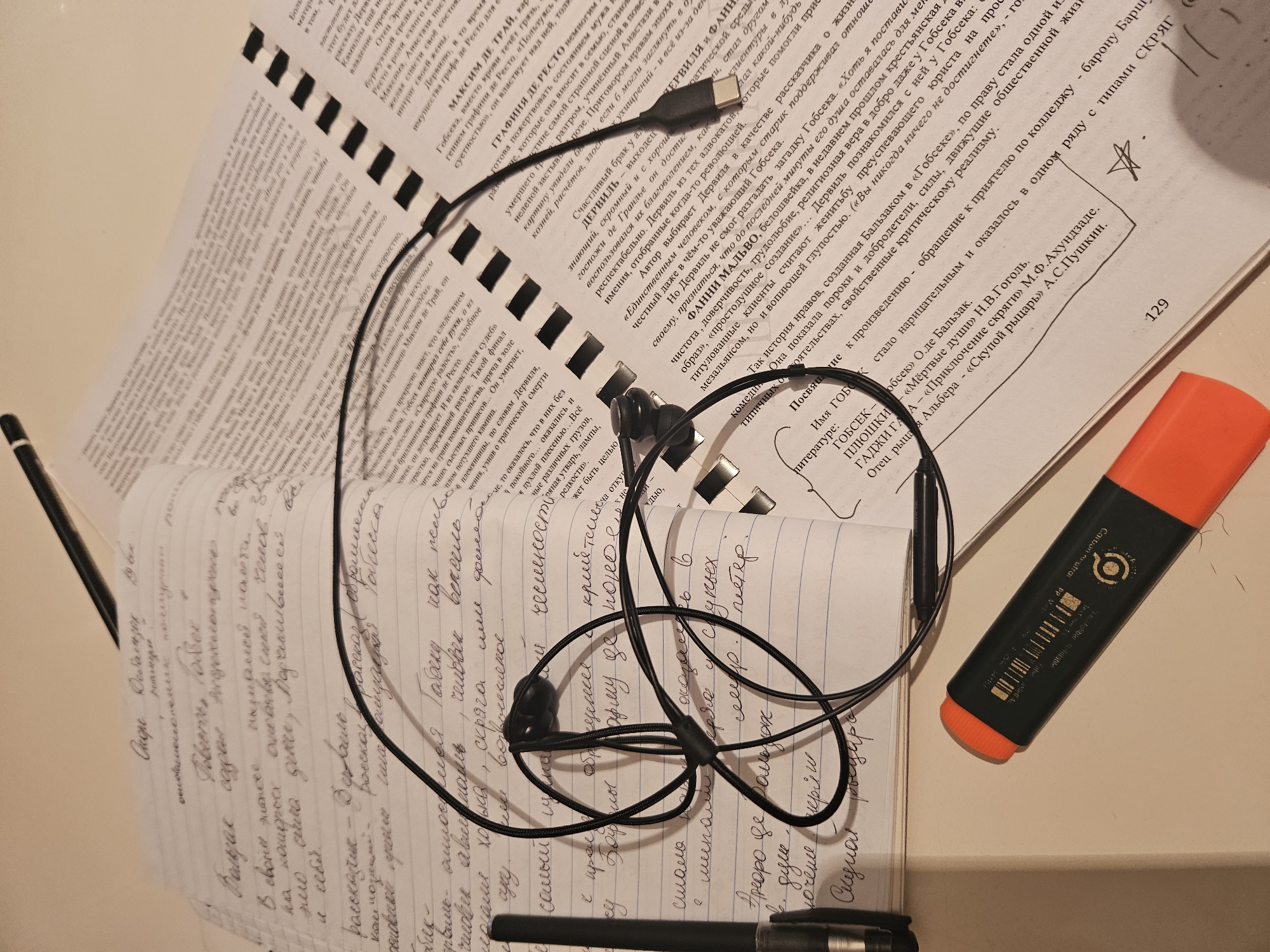 The image shows a cluttered desk with an open notebook, a printed book, a pair of black earbuds, and an orange marker. The notebook is filled with handwritten text in Russian. The printed book also contains Russian text. The earbuds are lying on top of the open notebook. The orange marker is positioned in the lower right corner of the image. The lighting is bright, suggesting an indoor environment. No people are visible in the image. The location is not specified in the image.
