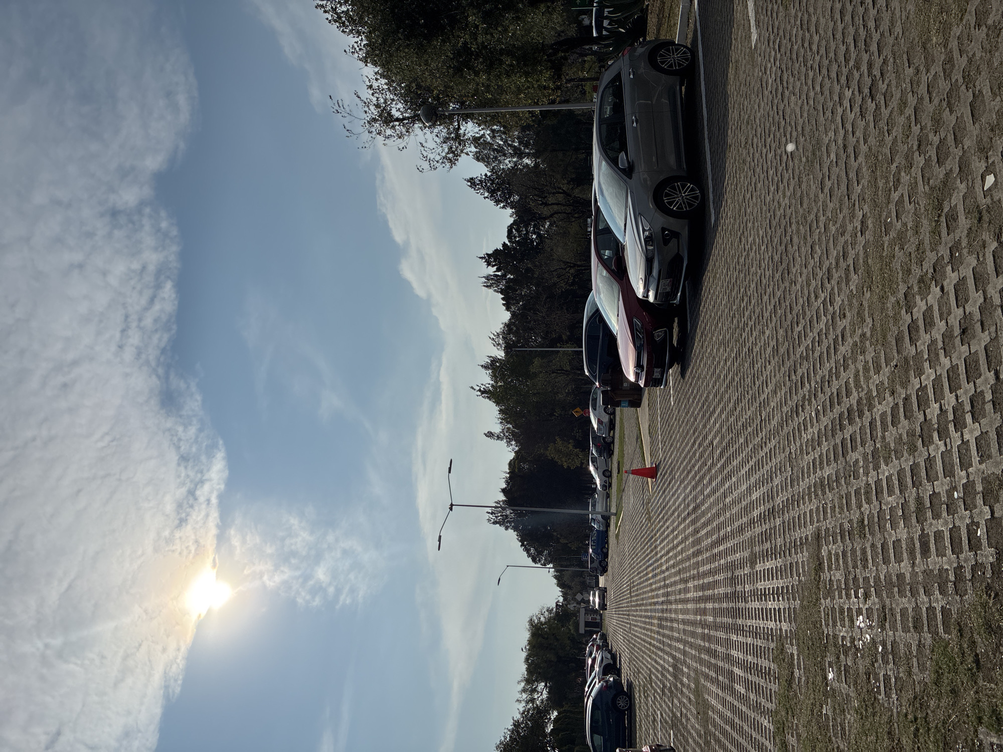 The image shows a parking lot in San Andrés Cholula, Mexico. The parking lot is paved with interlocking concrete blocks. A line of parked cars is visible on the left side of the frame. The sky is blue with clouds and the sun is visible. The trees are also visible. There is an orange cone placed.