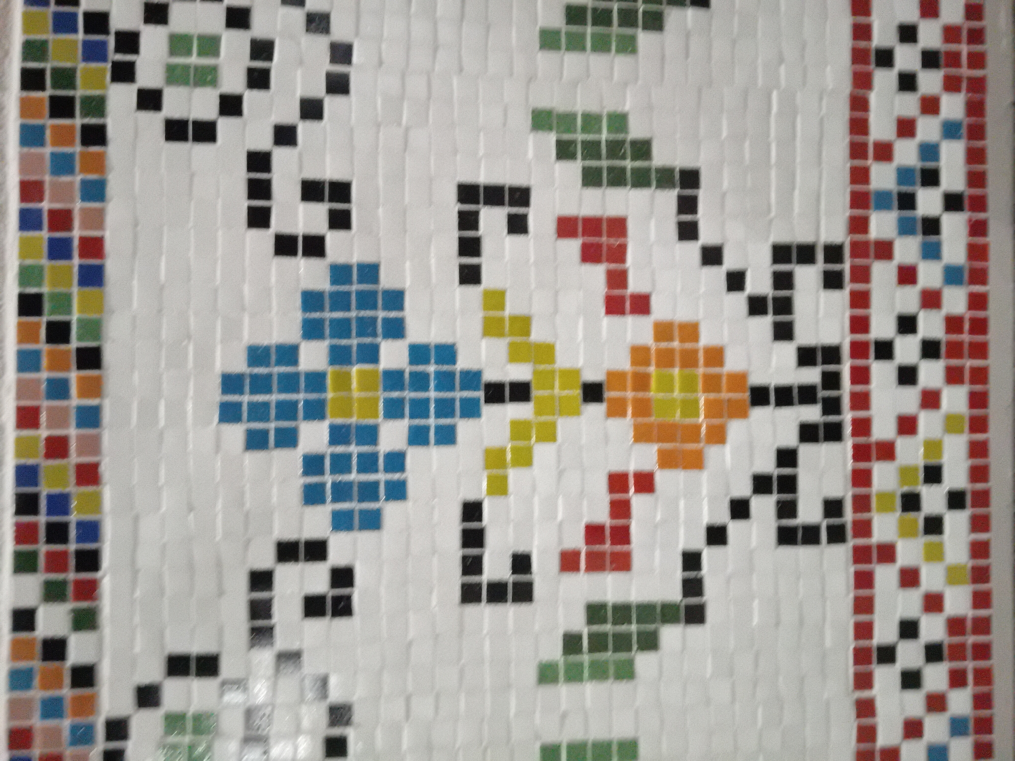 The image displays a close-up view of a vibrant mosaic artwork composed of small, square tiles. The background of the mosaic is predominantly white tiles, creating a light canvas for the colorful patterns.
In the central area, there are two distinct, stylized floral or abstract patterns. One pattern features blue and yellow tiles, while the other uses orange and red tiles. These central designs are connected by black tile elements that resemble stems or branches. Green tiles are strategically placed to suggest leaf-like forms, further enhancing the botanical aesthetic.
On the far left side of the artwork, a vertical border or stripe is visible. This border consists of rows of various colored tiles, including yellow, blue, black, green, orange, and red, arranged in an alternating fashion. On the right side, another intricate vertical border is present, characterized by a repeating pattern of red, white, black, blue, and yellow tiles.
The overall aesthetic is geometric and pixelated due to the uniform square shape of the tiles. The tiles appear to have a slightly glossy finish, and the image is well-lit, highlighting the individual colors and the precise arrangement of the mosaic pieces. There are no people, explicit activities, specific locations, or visible text in the image.