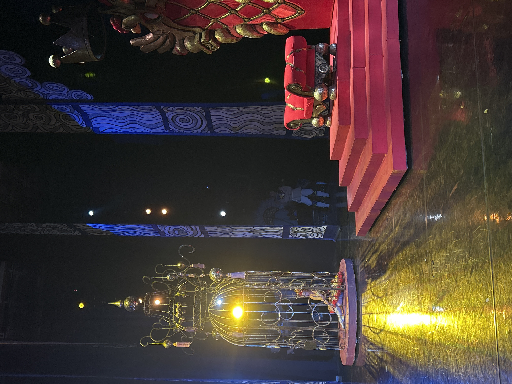 On a dark stage with reflective flooring, a theatrical scene is set. To the left, a woman in elaborate costume sits within a large, ornate, golden birdcage, illuminated by a bright yellow light. The cage itself is decorated with tassels and metallic spheres. In the center right of the stage, a set of plush red steps leads up to a red throne-like chair with gold accents. Behind the chair, a large, golden, ornate structure resembling a part of a crown or a royal headdress is visible. The background features tall, abstract pillars with blue and white swirling patterns. Scattered lighting from above creates beams and reflections on the stage floor.