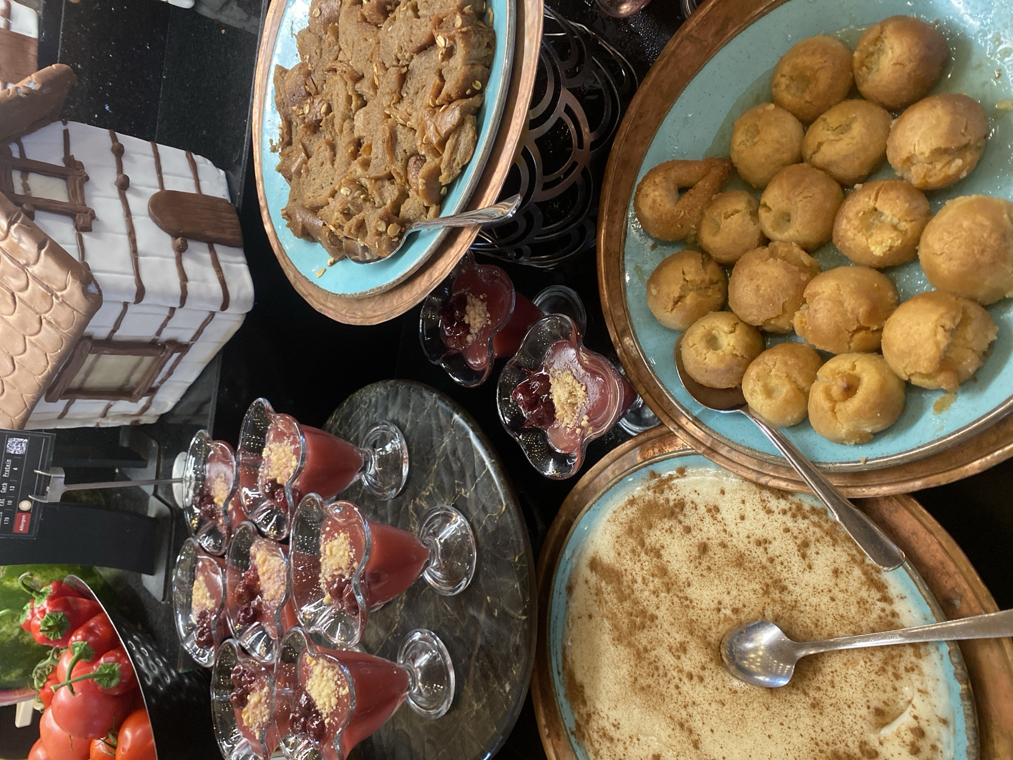 A buffet table displays a variety of desserts, including small glass cups filled with a red, jelly-like dessert topped with crumbled nuts and a cherry, a large platter of rice pudding dusted with cinnamon, and a turquoise platter filled with small, golden fried dough balls. To the right, a plate of what appears to be baked halva is served with a spoon. In the background, a gingerbread house decoration and a bowl of red peppers and tomatoes are visible. The scene appears to be a dessert spread at a hotel or restaurant in Aksu, Turkey. There are no people or animals visible in the image.