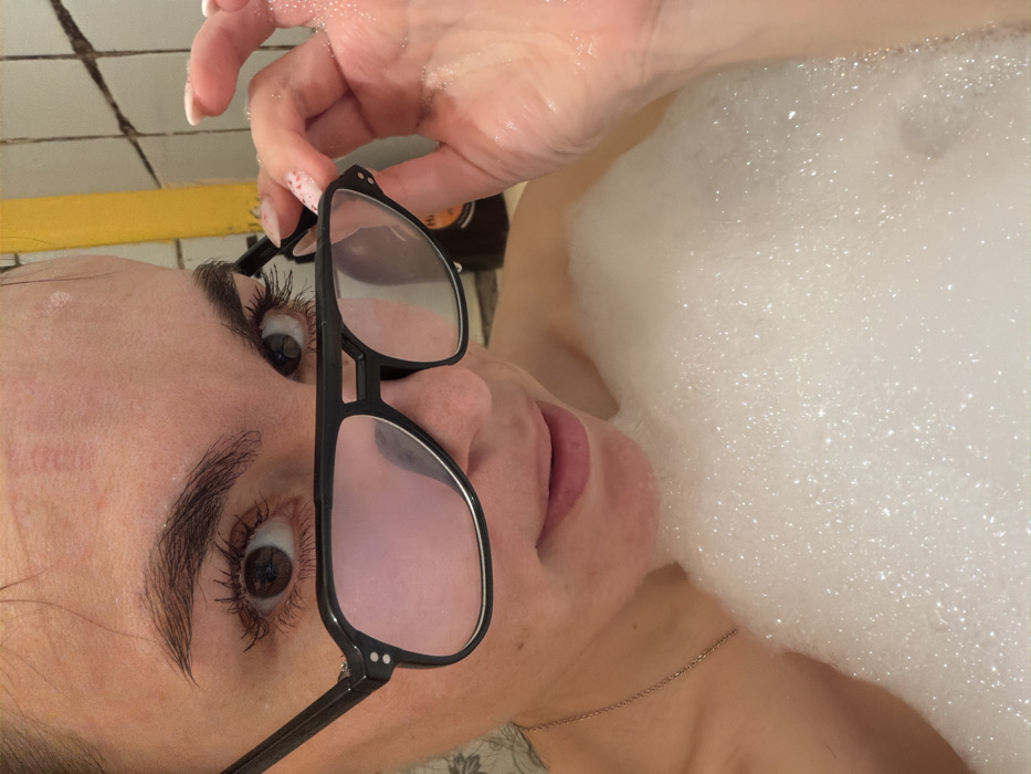 An adult female is depicted in a close-up, appearing submerged in a bath filled with white foam bubbles. Her head and shoulders are visible. She possesses wide, dark brown eyes with dark mascaraed lashes, and thick, well-groomed eyebrows. Water droplets are present on her forehead and upper cheeks, indicating wetness. Her lips are slightly parted. A thin metallic chain necklace is visible on the left side of her neck/chest. Her right hand, exhibiting light-colored fingernail polish and small adhering bubbles, holds a pair of black-framed, retro-style glasses with subtly pink-tinted lenses directly in front of her eyes. The subject gazes directly into the camera. The background consists of light-colored square wall tiles with dark grout lines, a vertical yellow fixture, and a dark product bottle. The environment is consistent with a private bathroom. The specific address or precise location within Izhevsk, Russia, cannot be determined from the visual evidence.