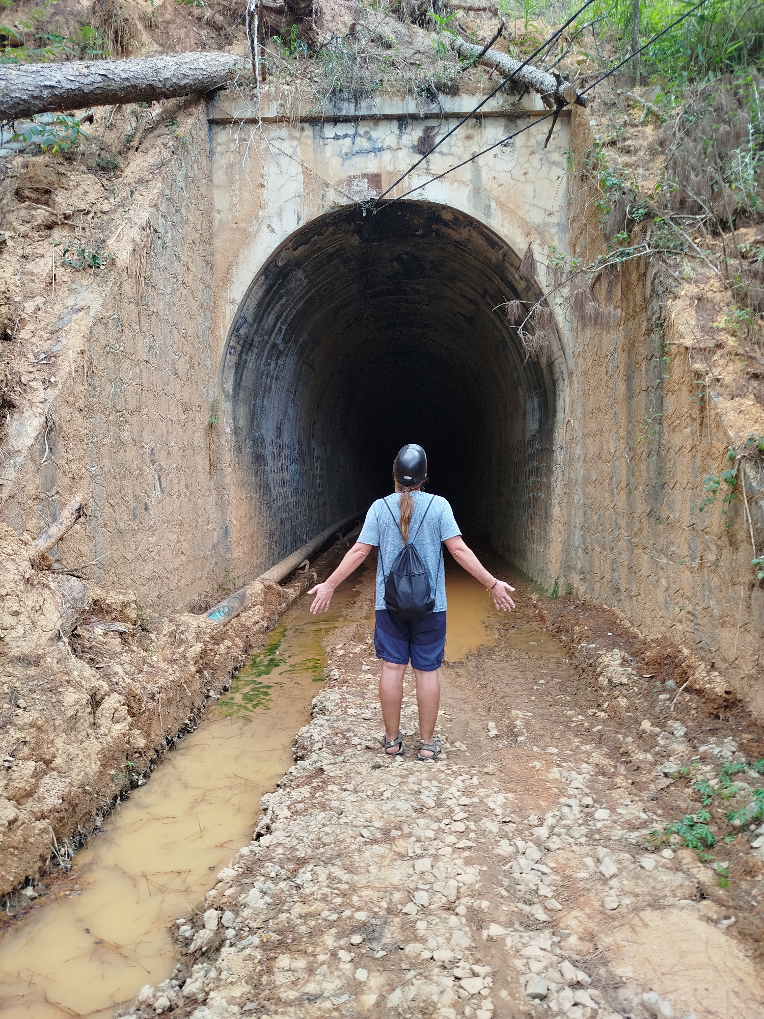 A middle-aged person stands facing the entrance of a dark tunnel. The person is wearing a light blue t-shirt, dark blue shorts, sandals, a black helmet, and a black backpack. Their arms are outstretched at their sides. The tunnel entrance is constructed of stone and concrete, with vegetation and fallen trees visible on the upper edges. The ground leading to the tunnel is uneven and muddy, with a water-filled ditch along the left side. The walls of the tunnel entrance are covered in dirt. Overhead, black wires run along the top of the tunnel. The location is in Đa Thọ, Vietnam.
