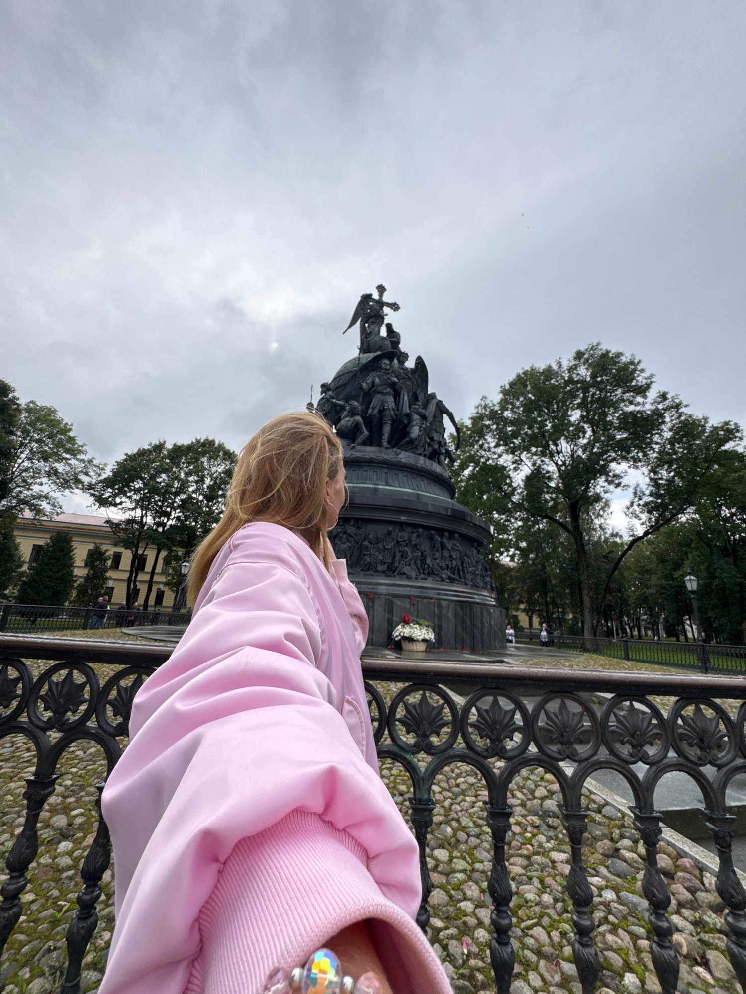 In Veliky Novgorod, Russia, a blonde-haired woman in a pink jacket stands facing a large monument on an overcast day. The monument, a dark-colored statue on a substantial base, features numerous figures and a cross at its apex. In the foreground, a decorative black metal fence with intricate patterns separates the viewer from a cobbled pathway. Behind the monument, trees and a building with yellow walls are visible. The sky is filled with clouds.