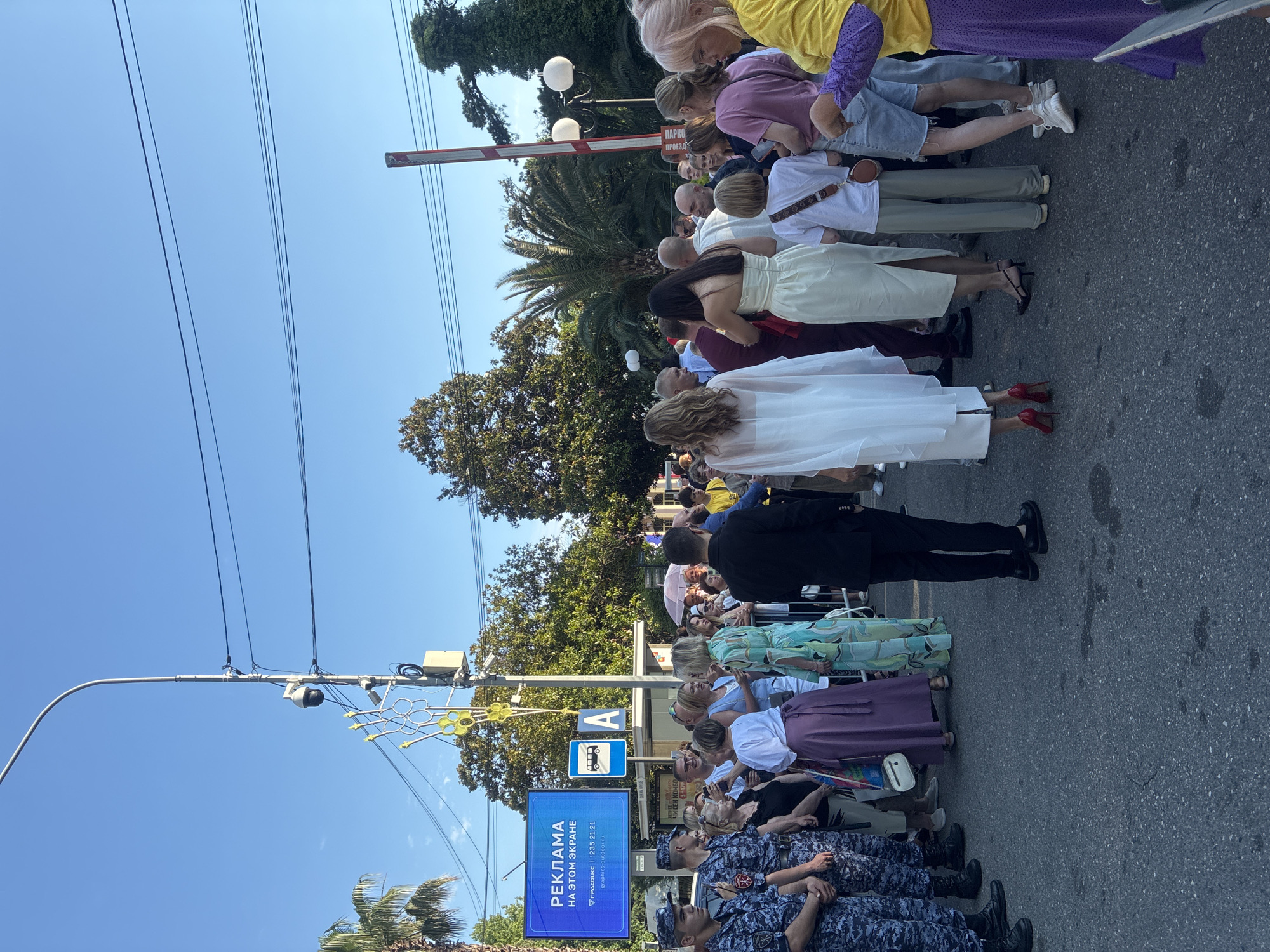 In Sochi, Russia, on a clear day, a crowd of people gathers on a paved street. Two individuals in dark blue police uniforms stand on the left, facing forward. A large blue digital billboard on the left displays the text "РЕКЛАМА НА ЭТОМ ЭКРАНЕ" which translates to "ADVERTISEMENT ON THIS SCREEN." Below this, smaller text reads "ГРАФИСС | 235 21 21" and "graphics-outdoor.ru". Next to the billboard is a blue bus stop sign with a white bus icon and the letter "A." In the background, palm trees and other foliage are visible, along with streetlights and power lines. A red and white barrier gate is partially visible on the right side of the image. The people in the crowd are diverse in age and dress, with some individuals wearing formal attire and others in casual clothing. Some are looking towards the left of the frame, seemingly at something of interest. The overall scene suggests an outdoor event or gathering in a public space.