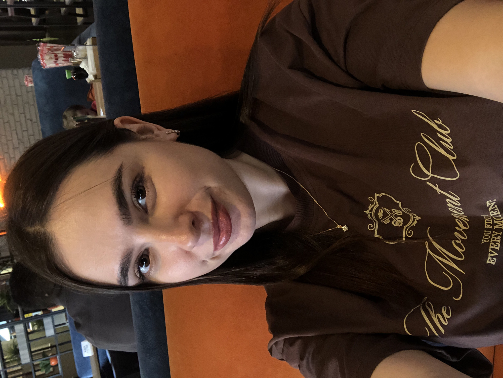 The image shows a woman with dark hair, wearing a brown t-shirt with gold lettering and a small diamond pendant necklace. She is sitting in what appears to be a restaurant or cafe with an orange seat visible in the foreground. In the background, there are blurred figures and seating arrangements, including a blue couch and brick wall. The overall impression is a casual selfie taken indoors. The specific location in Izhevsk, Russia, cannot be determined from the visual information provided.