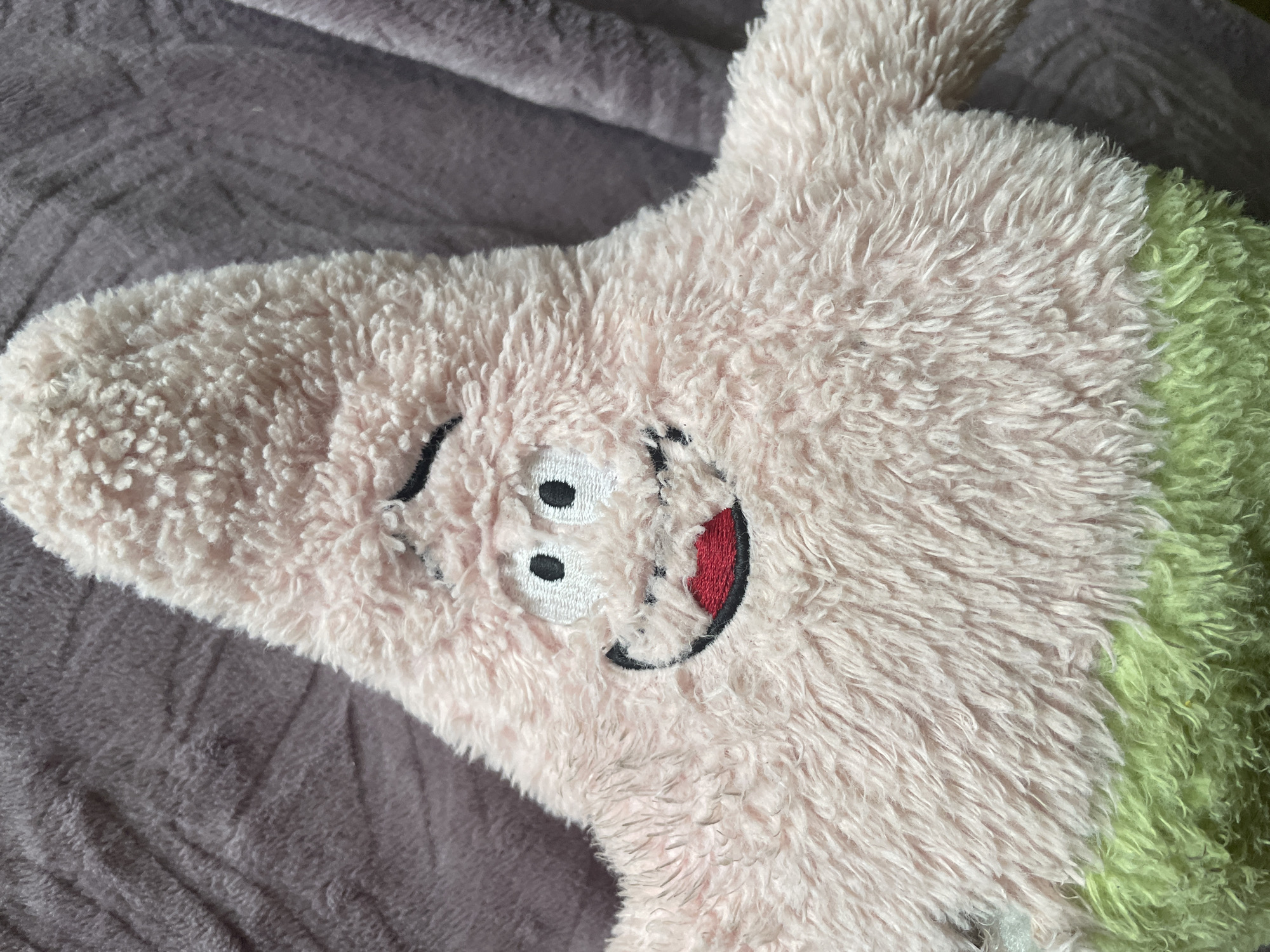 A close-up, overhead shot shows a pink, plush stuffed toy with a cartoon face. The toy is lying on a textured, purple fabric surface. The face of the toy has large, white, circular eyes with black pupils, a wide, red mouth outlined in black, and black eyebrows. The plush fabric of the toy is light pink and appears soft and fluffy. A portion of the toy is a greenish color. The background fabric is a dull purple with visible wrinkles and a soft texture, suggesting it may be a blanket or a rug. The lighting is even, with no harsh shadows, indicating it might be taken indoors with diffused light. The location is Unknown, Unknown.