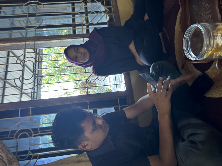 The scene is an interior room, likely a residence, in Pante, Indonesia. Two individuals, an adult male and an adult female, are seated on a patterned floor covering. The male, positioned on the left, wears a dark short-sleeved collared shirt and dark trousers. He is observed looking downward, engaged with a dark-colored smartphone held in both hands. The female, positioned on the right, wears a dark long-sleeved garment, dark trousers, and a dark purple hijab. She is observed looking directly forward, towards the camera's perspective, with a neutral expression. Behind both subjects is a large multi-pane window with ornate, light-colored metal grilles, through which bright daylight illuminates green foliage and trees outdoors. In the bottom right foreground, a clear glass jar containing light-colored, possibly edible, irregular solids is positioned on a decorative wooden tray.