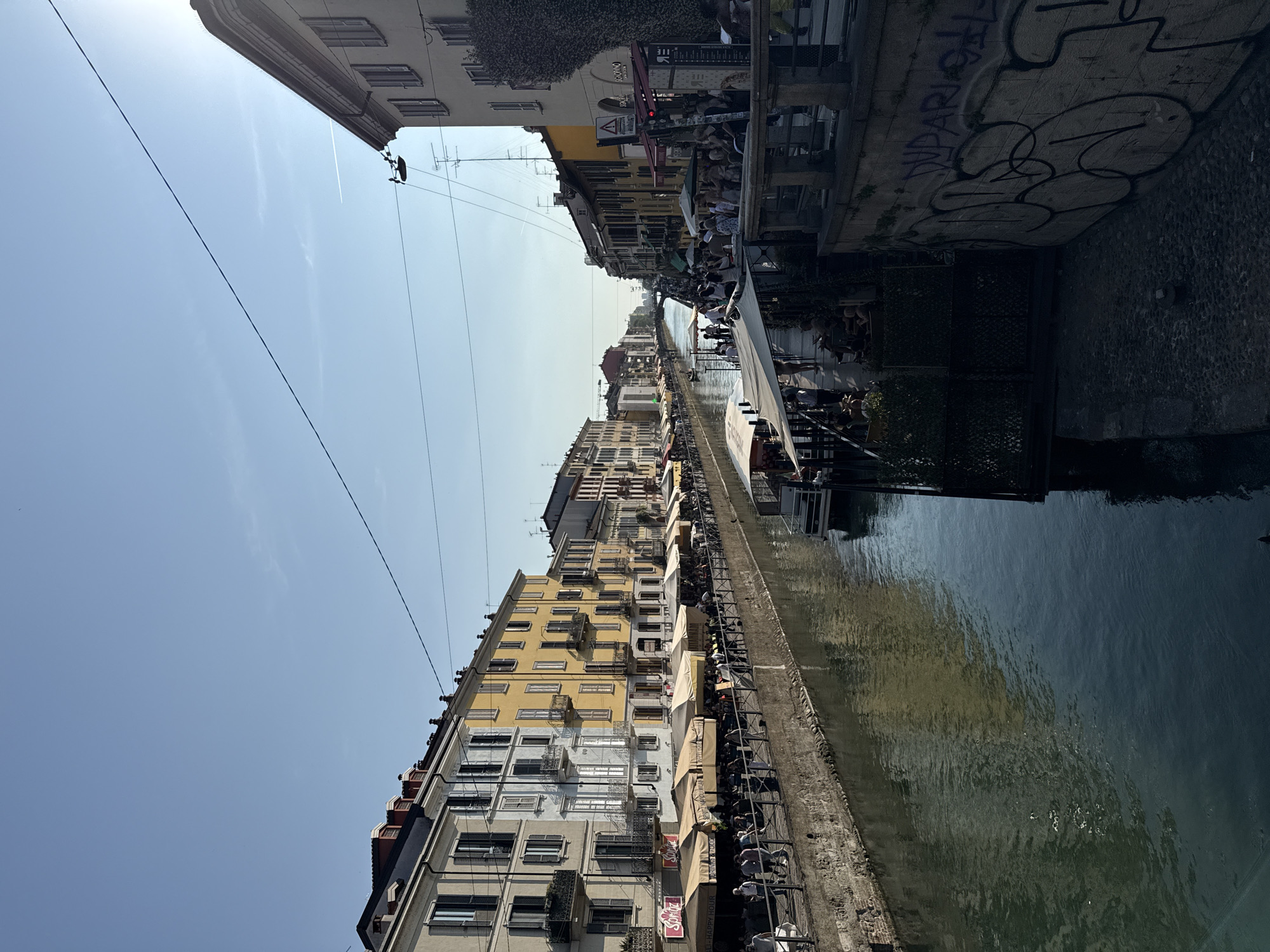 The image captures a canal in Milan, Italy, lined by multi-story buildings on both sides. The canal water reflects the buildings and the sky above. There are people along the canal, many seated at tables under awnings, likely patrons of cafes or restaurants. Graffiti marks a section of the canal wall. Power lines stretch across the sky above the buildings. The buildings have a variety of architectural styles and colors, with many windows and balconies visible. The environment appears to be urban and bustling, with a focus on leisure and social activity.