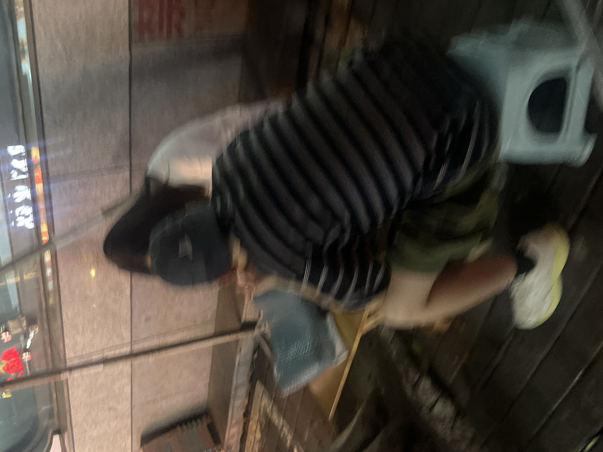 Two individuals are seated on a wooden platform at night, presumably in Yuzhong District, China. The individual in the foreground, wearing a dark striped shirt and a cap, is seated on a light blue stool. Their legs, clad in shorts and white sneakers with yellow soles, are visible. A teal bag rests on a wooden surface in front of them. Behind this person, another individual with long dark hair is seated, wearing a white top. The background shows a textured wall with some illuminated signage above it. The overall scene is dimly lit and the image quality is blurry, indicating motion or low light conditions.