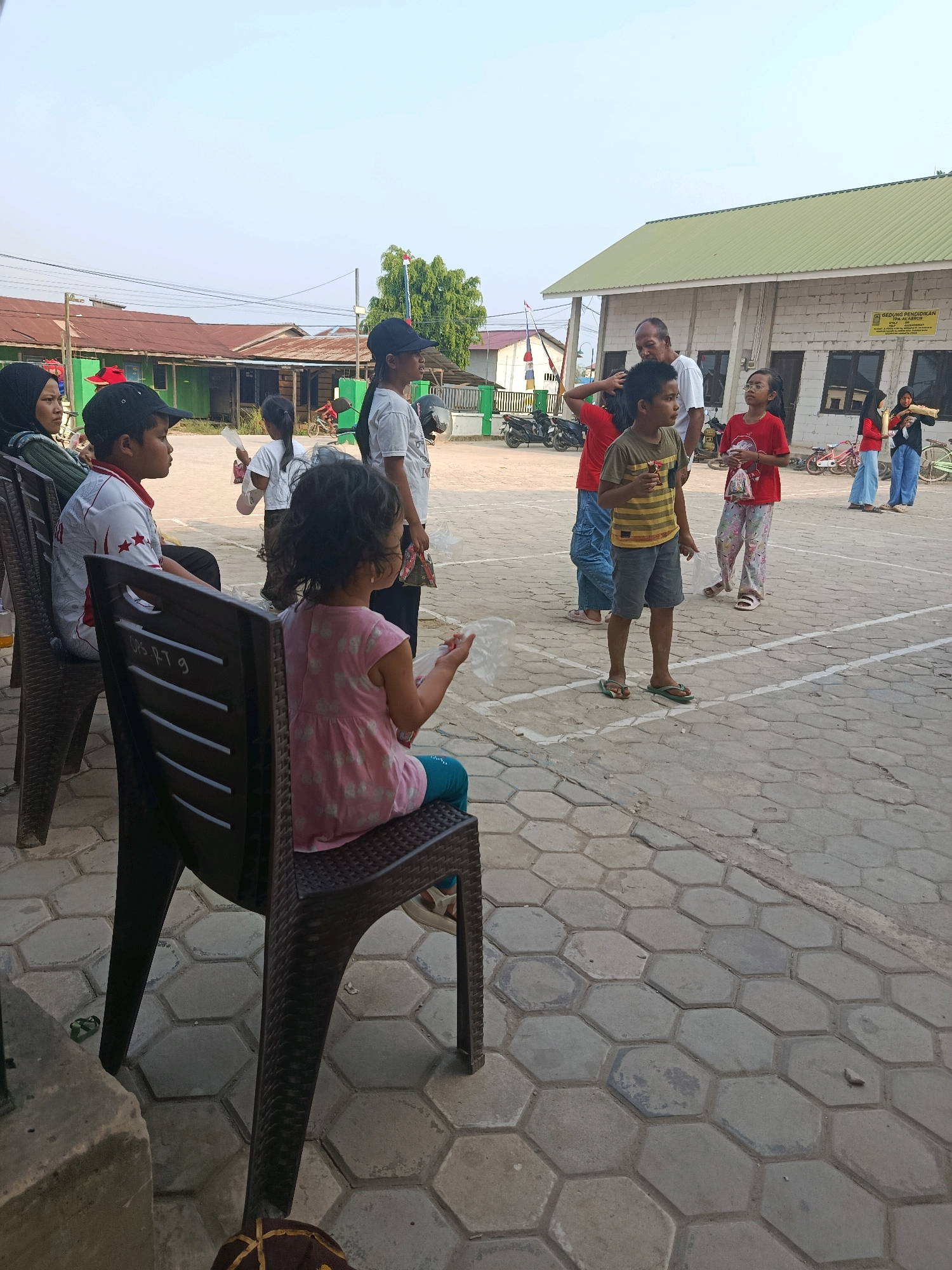 This image depicts an outdoor scene in Swarga Bara, Indonesia, likely a community area or school grounds, given the presence of a building labeled "GEDUNG PENDIDIKAN TPA AL-ABROR". The foreground features several individuals, primarily children and some adults, gathered on a paved area with a hexagonal tile pattern.
In the immediate foreground, a young girl with dark curly hair is seated in a dark plastic chair, facing to the right, wearing a pink sleeveless dress with a white floral pattern and teal leggings. She appears to be holding a clear plastic bag. To her left and slightly behind her, a young boy with dark hair and wearing a black baseball cap is also seated in a dark plastic chair, looking to his right. He is wearing a white shirt with red and black accents and possibly holding a snack.
Further back, a group of children and adults are standing. A boy in a yellow and black striped t-shirt and gray shorts is looking towards the right. A man in a white t-shirt is standing behind him. Several other children are visible, some holding clear plastic bags. In the background, there is a building with a light green roof and white concrete block walls. There are also some smaller buildings with corrugated metal roofs, parked motorcycles, and trees. The sky is clear and bright, suggesting daytime. The overall atmosphere appears to be relaxed and communal.