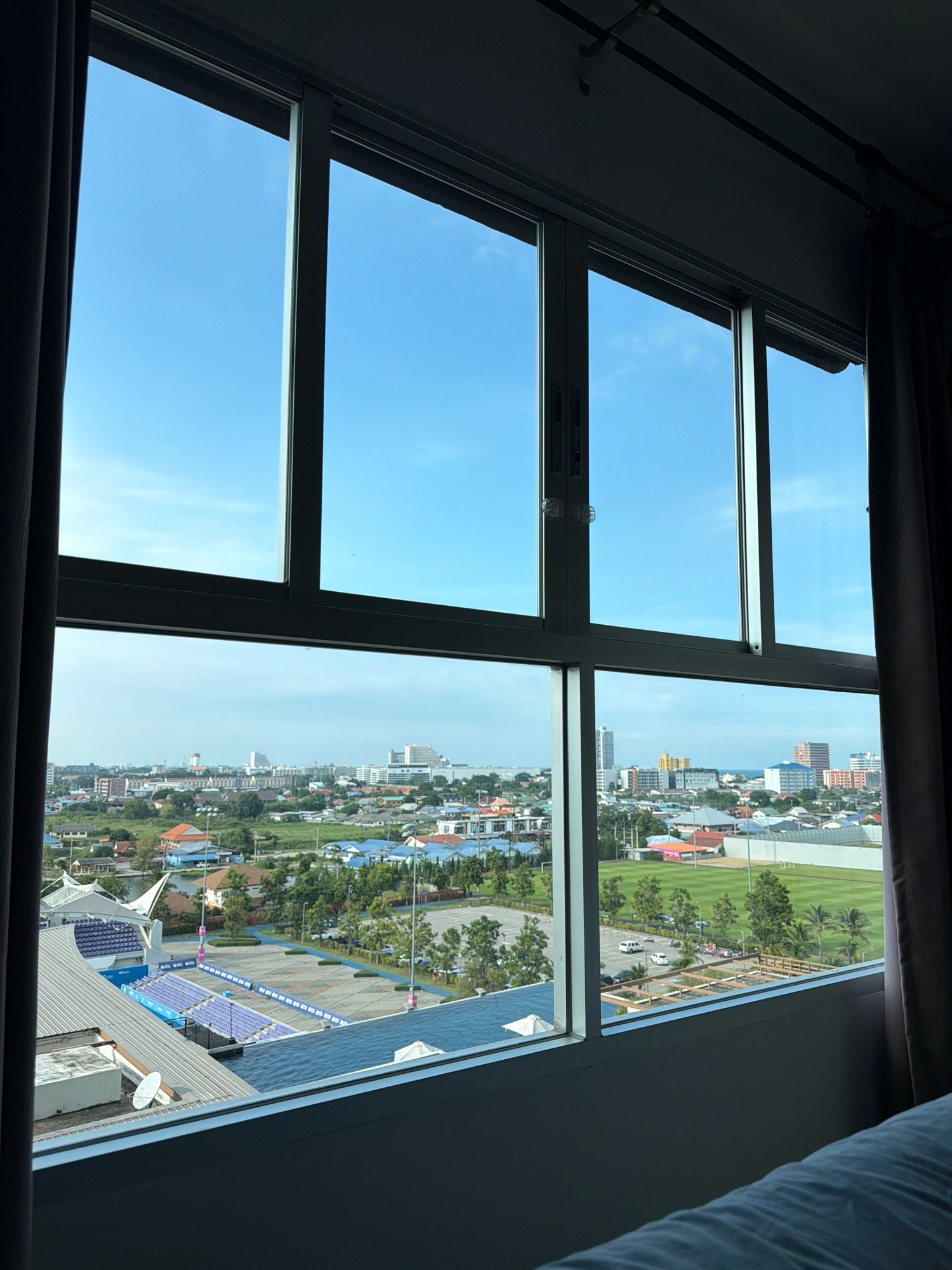 The image shows a view from a window looking out over the city of Hua Hin, Thailand. The foreground is dominated by a large window frame and curtains on either side. Through the window, a cityscape unfolds with a clear blue sky overhead.
In the mid-ground, a sports stadium with blue seating is visible, along with a large paved area and some trees. Beyond the stadium, the city extends outwards with numerous buildings of varying heights and architectural styles, including some taller structures that appear to be hotels or apartment buildings. Green spaces, including what looks like a sports field with manicured grass, are interspersed among the buildings. Roads with a few cars are also visible. In the distance, a hazy horizon suggests the presence of the ocean. The overall impression is of a coastal city with a mix of residential, commercial, and recreational areas.