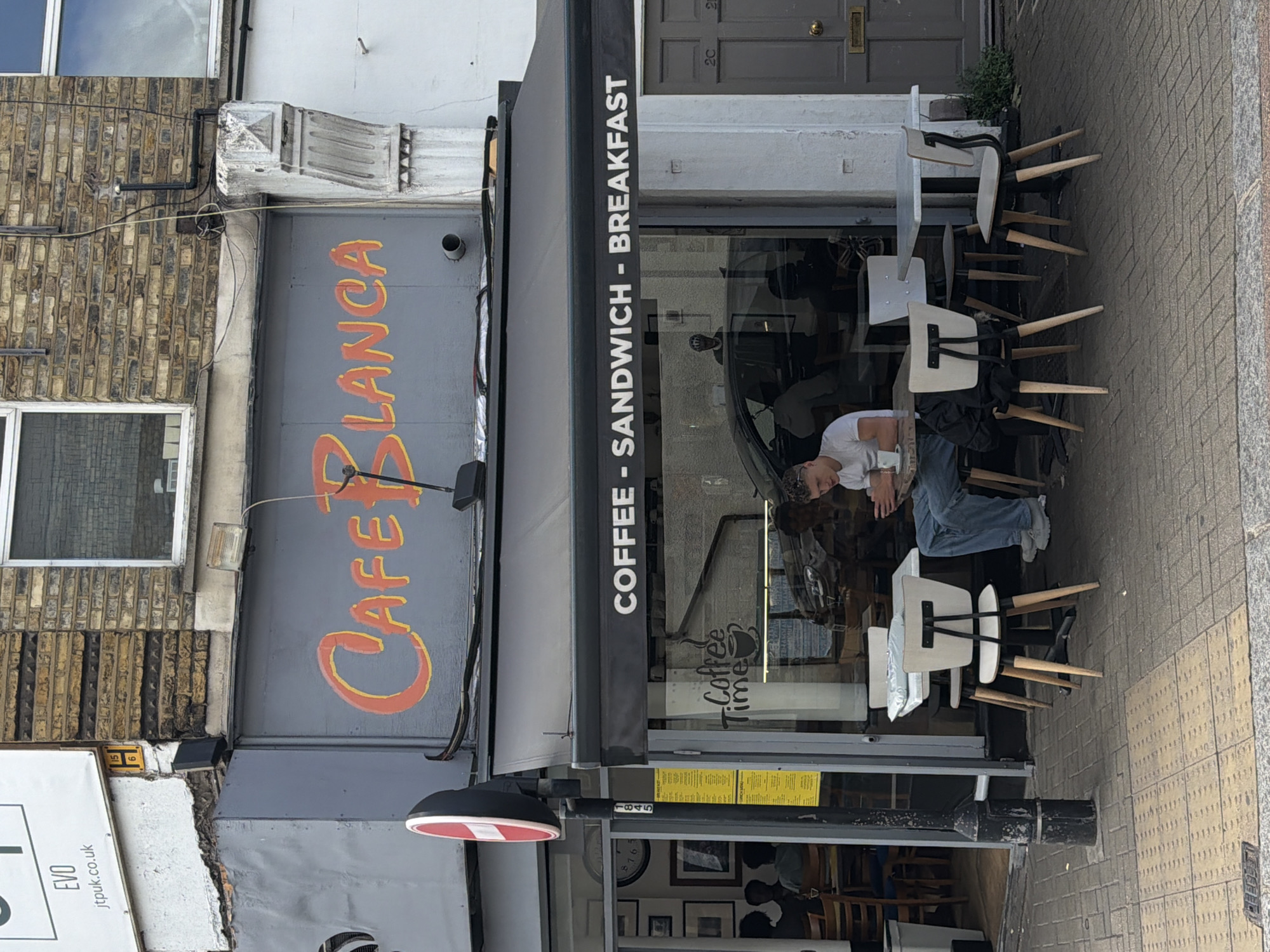 A street view outside a cafe shows a man sitting at a table, looking at his phone. The cafe, named "Cafe BLANCA," has a sign above its entrance that reads "COFFEE - SANDWICH - BREAKFAST." The cafe is located in London, United Kingdom. The man is wearing a white t-shirt and blue jeans, and he is seated at a table with chairs. There are other tables and chairs outside the cafe, and a black car is parked in front of the building. The street is paved with bricks, and there is a sidewalk with a tactile paving.