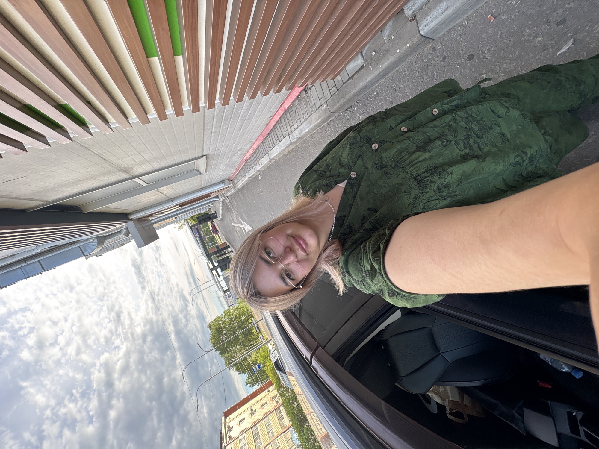 A young woman with blonde hair and glasses takes a selfie outdoors. She is wearing a green patterned dress and a pearl necklace. The woman is standing next to a black car. The background shows a street with buildings, trees, and a cloudy sky. The building on the right has a modern facade with horizontal wooden and green panels. In the distance, a multi-story apartment building is visible. The overall scene appears to be in an urban environment, likely in Birobidzhan, Russia, given the context.